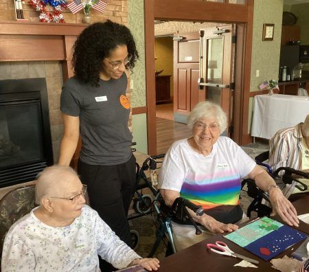 A staff member assists two older adults at a table with an arts and crafts project in a community room.