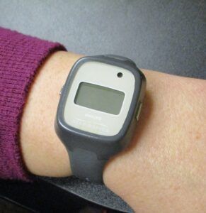 A gray, rectangular medical alert monitor worn on a person's wrist, featuring a blank digital display screen.