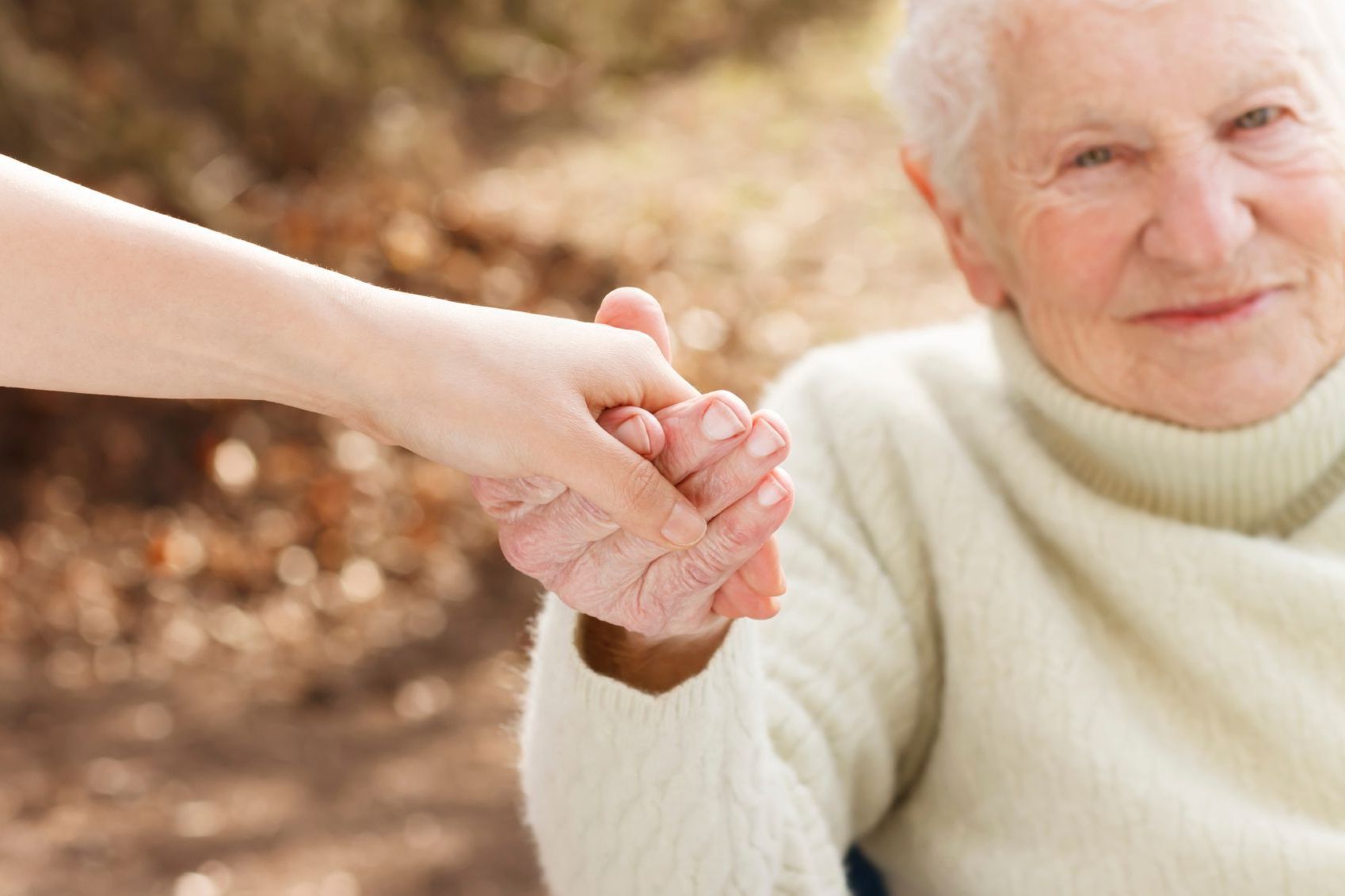 A person holding the wrinkled hand of another person in a gesture of support, with a blurred outdoor background.