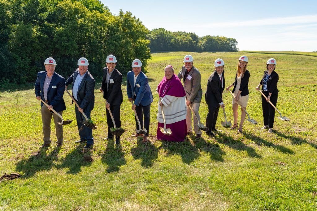 A group of people in hard hats and formal attire participate in a ceremonial groundbreaking in a grassy field.