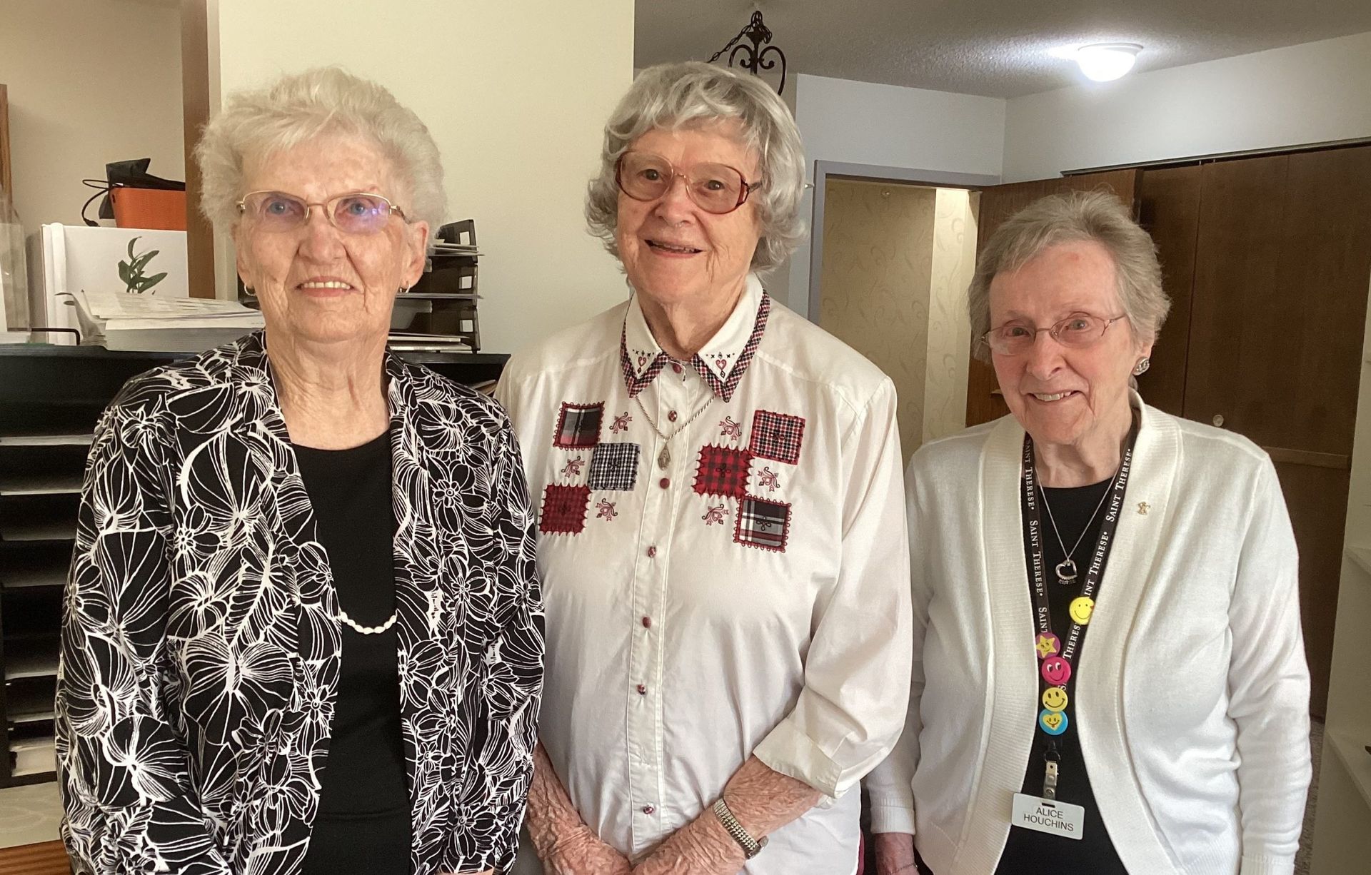 Three individuals stand together indoors, smiling while wearing casual clothing and glasses in a brightly lit room.