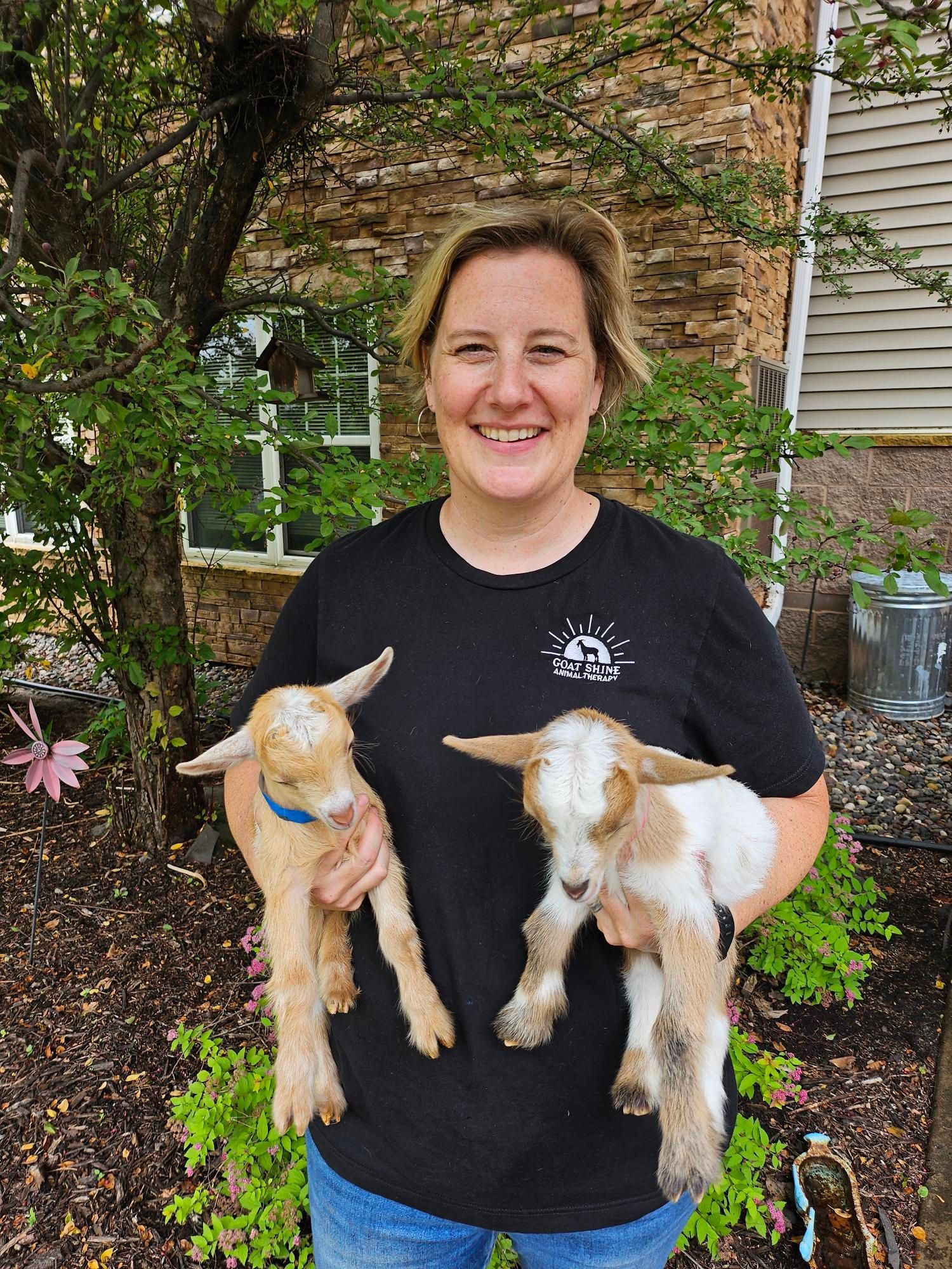 A person smiling while holding a small tan goat in each arm, standing outdoors in front of trees and a building.