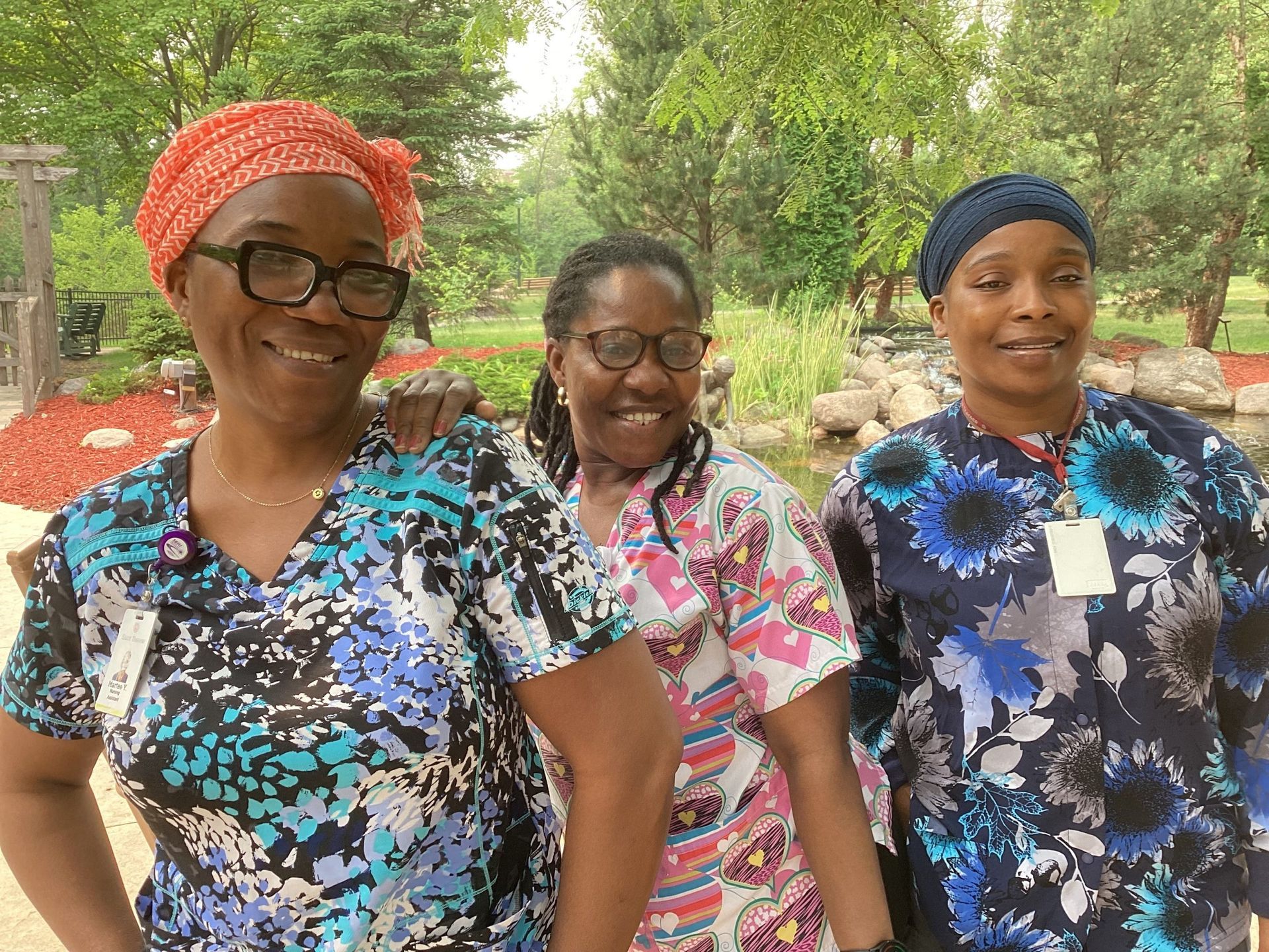 Three smiling people in patterned scrubs stand outdoors in a park-like setting with trees and a stone-lined pond.