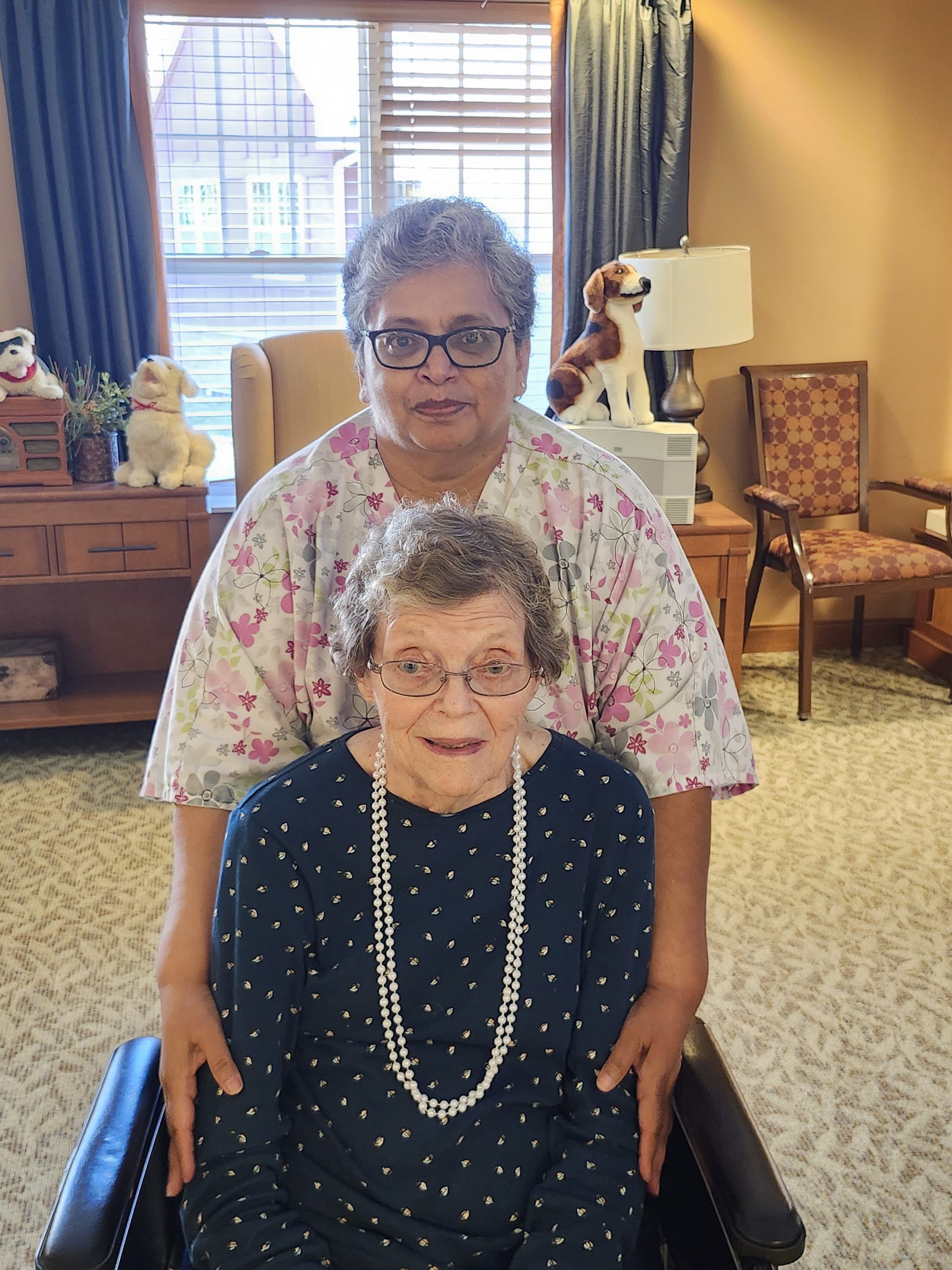 Two people pose together in a room; one stands behind the other, who sits in a chair wearing a long, beaded necklace.