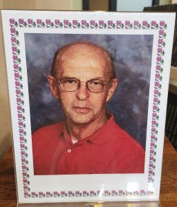 A framed portrait of a person wearing glasses and a red collared shirt, set against a blurred gray background.