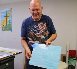 A person in a flag t-shirt holds up a blue painted canvas in a studio with art supplies and a wall decoration.
