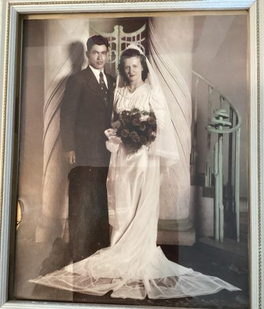 A vintage, framed wedding photograph showing a couple standing indoors near a staircase, holding a bouquet of flowers.