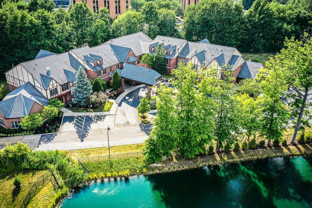 An aerial view of a multi-story building with a gray roof, surrounded by green trees and a small, bright blue pond.