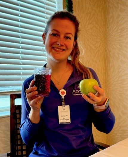 A smiling person in a blue zip-up holding a glass of dark liquid and a green apple indoors near window blinds.