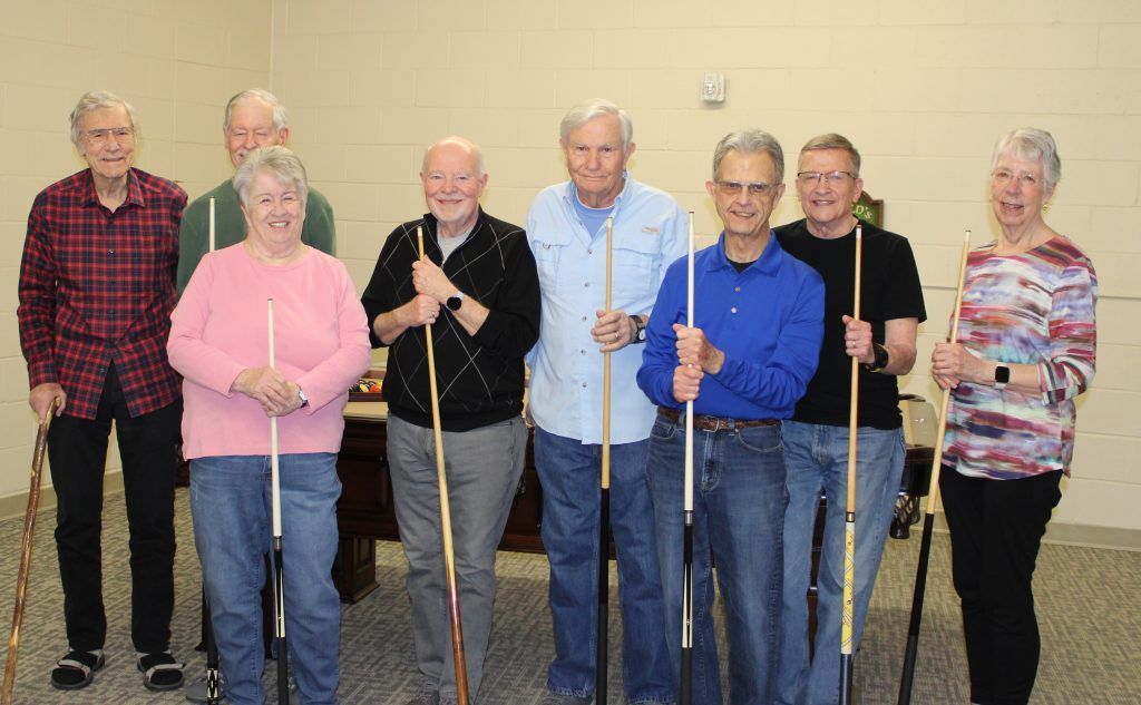 A group of people standing in a row, holding pool cues in a brightly lit room.