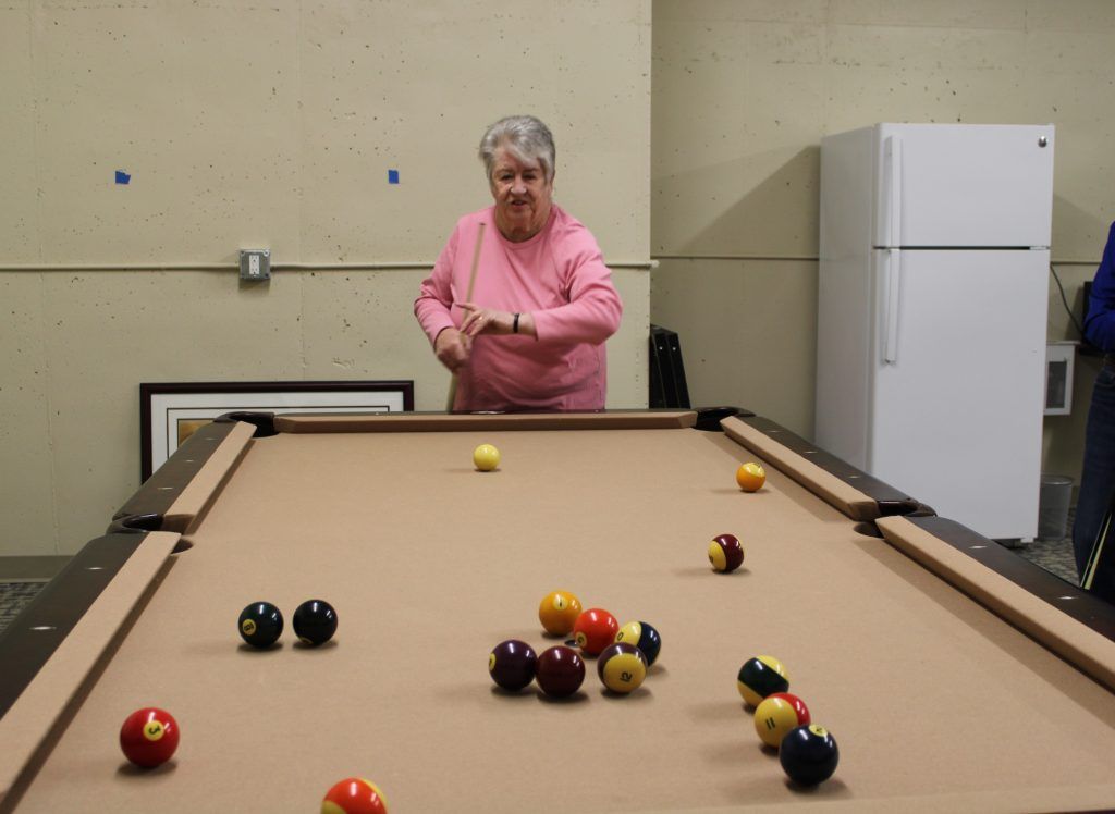 A person in a pink long-sleeved shirt prepares to hit a yellow ball on a tan pool table with scattered billiard balls.