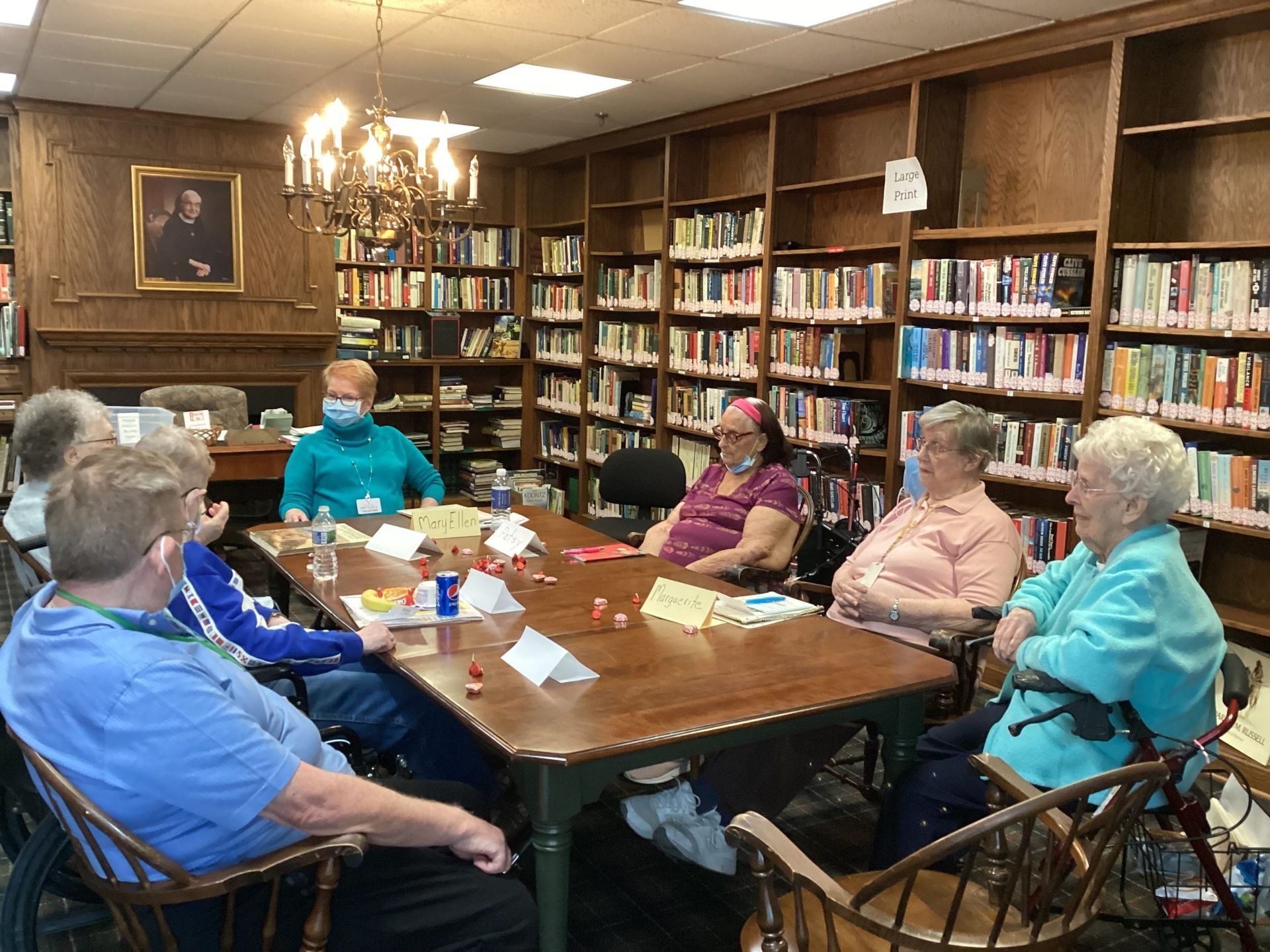 A group of people sit around a long wooden table in a library with floor-to-ceiling bookshelves, engaged in discussion.