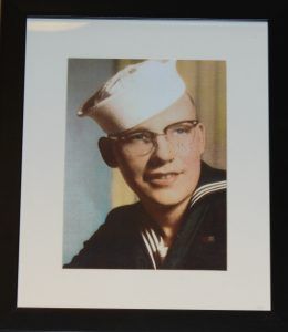 A framed portrait of a person in a US Navy uniform and white service cap, wearing glasses and smiling slightly.