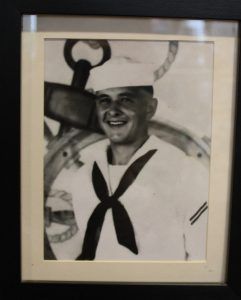 A black-and-white portrait of a sailor in uniform smiling in front of a ship’s anchor and wheel.
