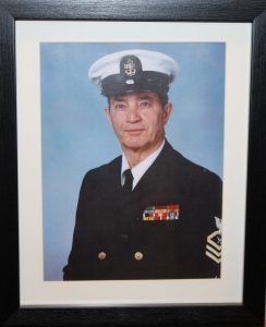 A formal portrait of a United States Navy Sailor in a dress blue uniform with a white service hat, framed on a wall.