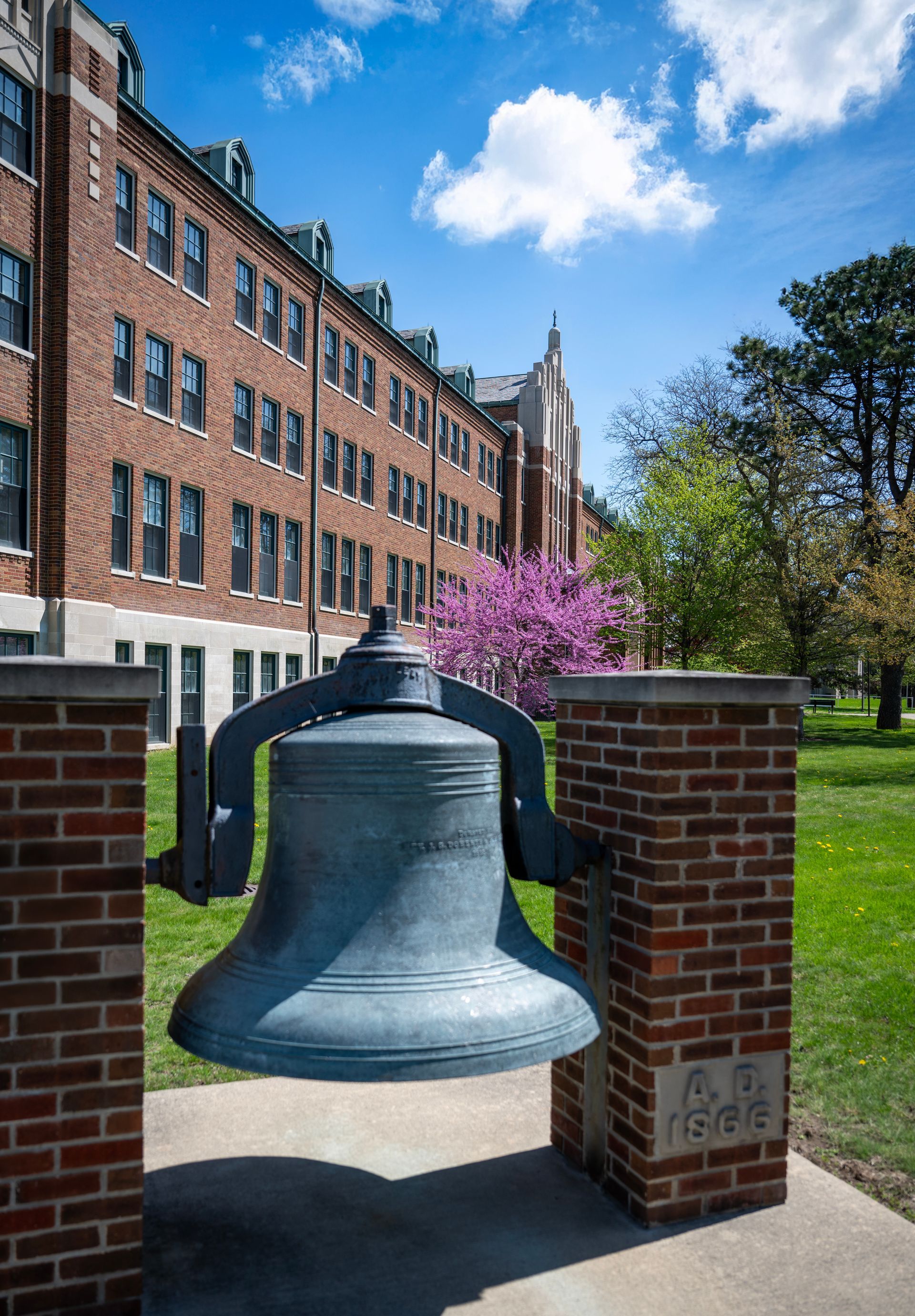 Large bronze bell between brick pillars on a campus lawn with buildings and trees in the background