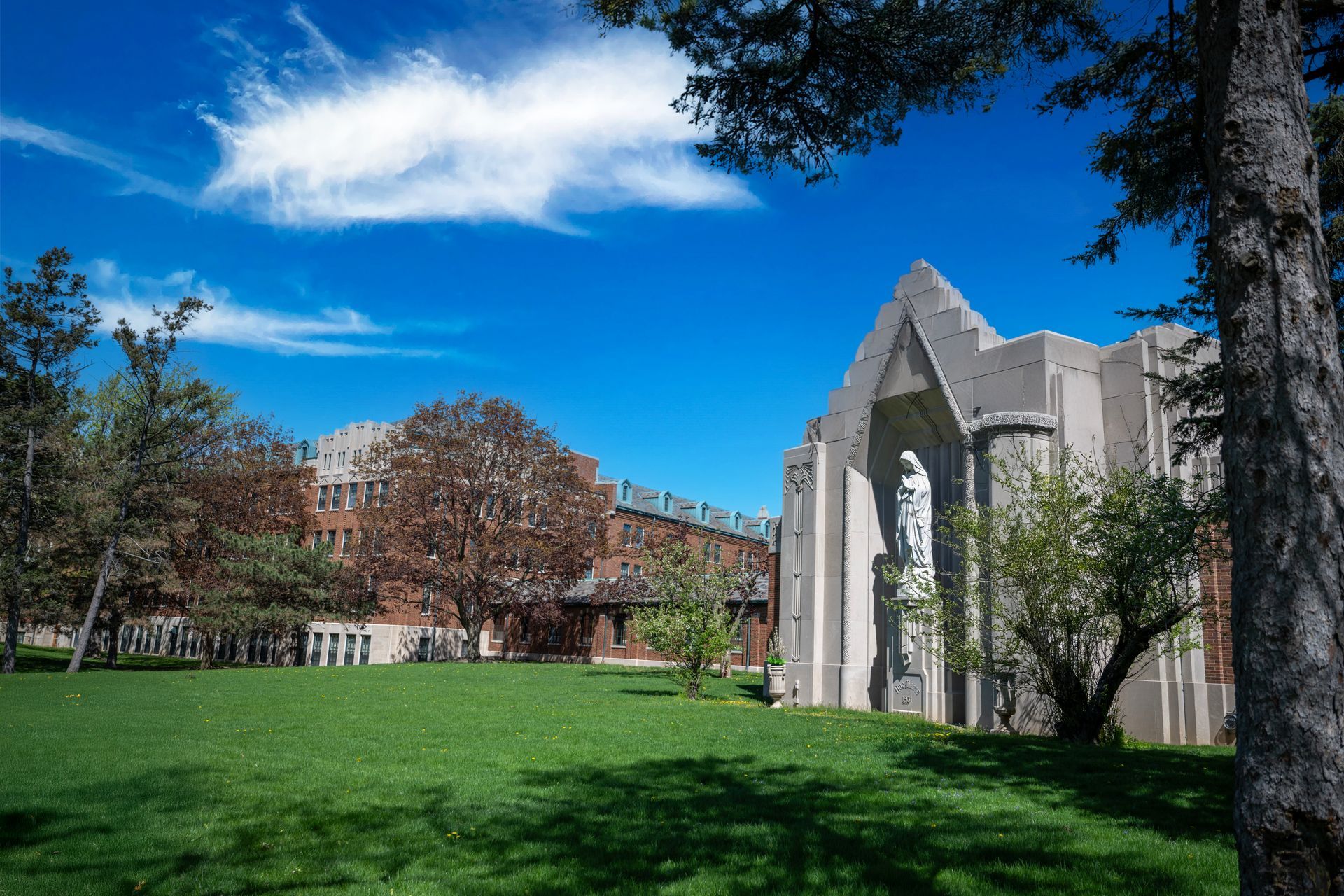 Campus lawn with a white stone building, green grass, trees, and a bright blue sky