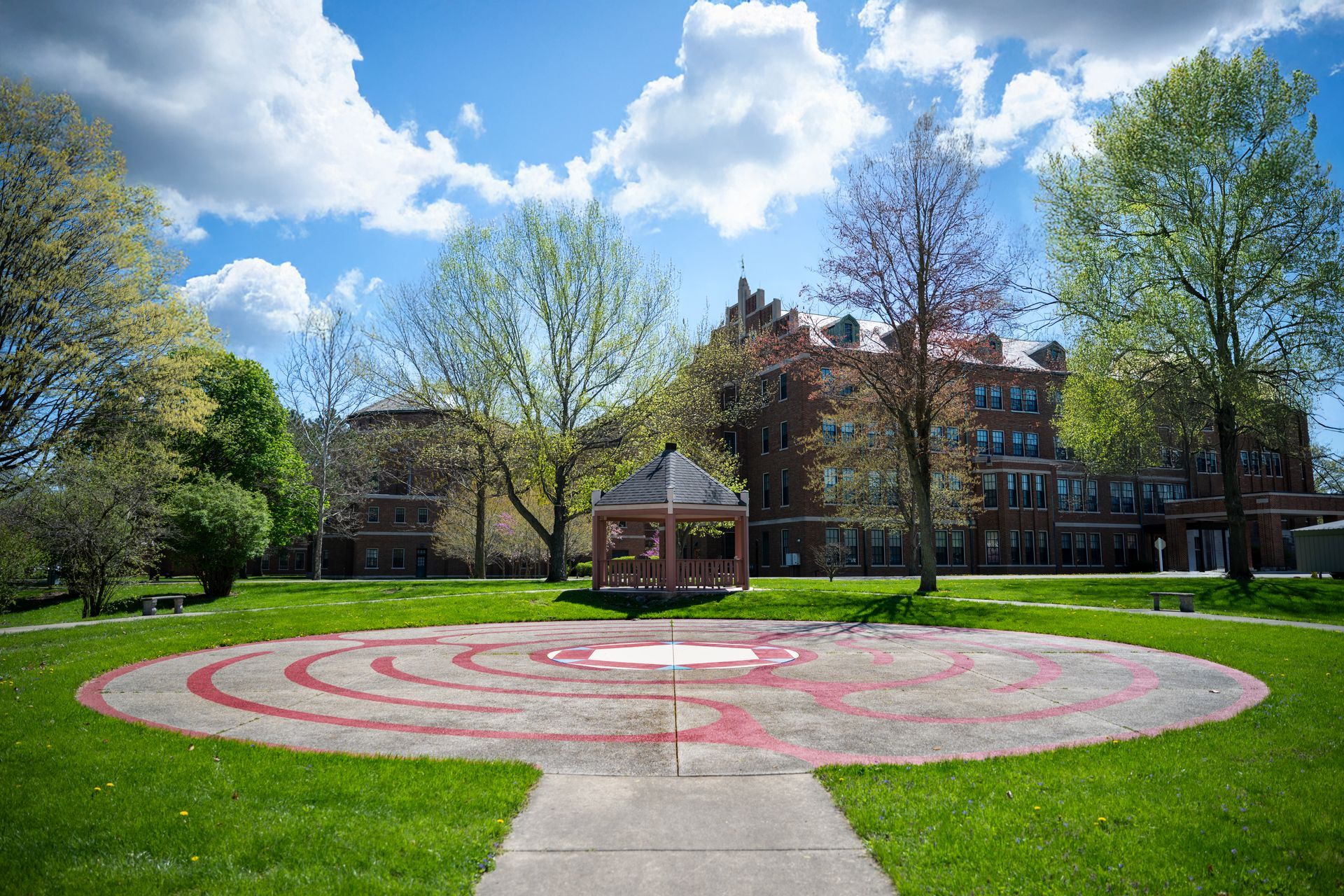 Park lawn with a red-and-white circular pattern, gazebo, trees, and buildings under a partly cloudy sky