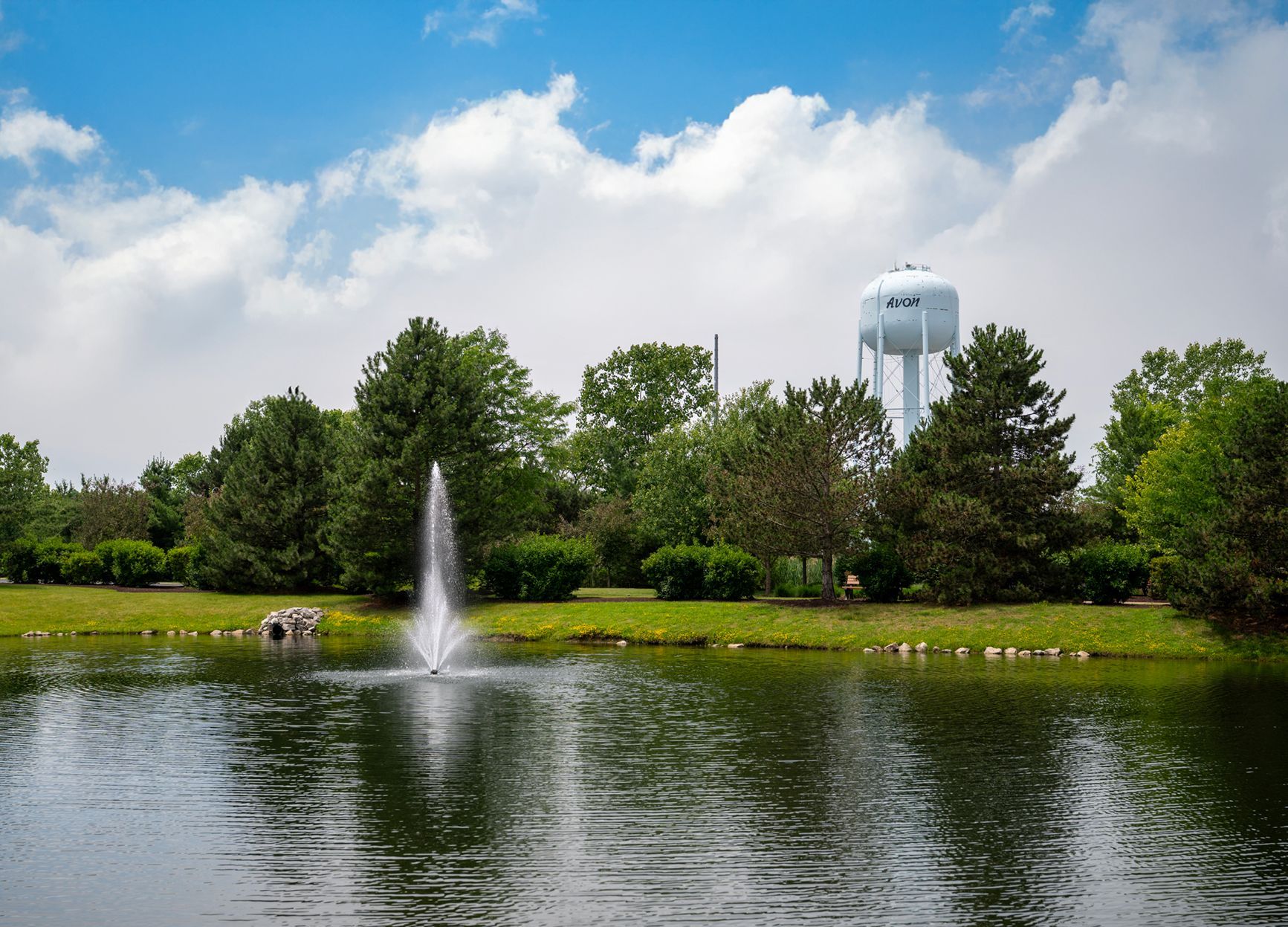 A water fountain sprays in a lake surrounded by lush green trees under a bright blue sky with a white water tower behind.