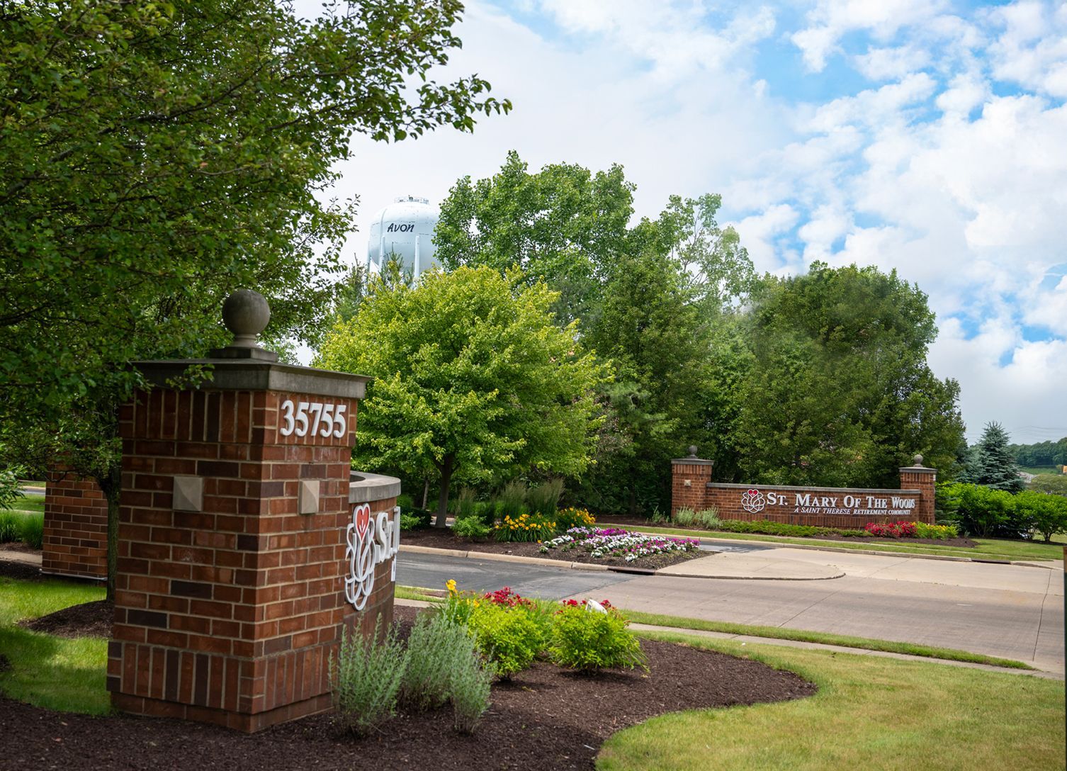 A brick neighborhood entrance sign with address 35755, lush greenery, and a water tower in the distance.