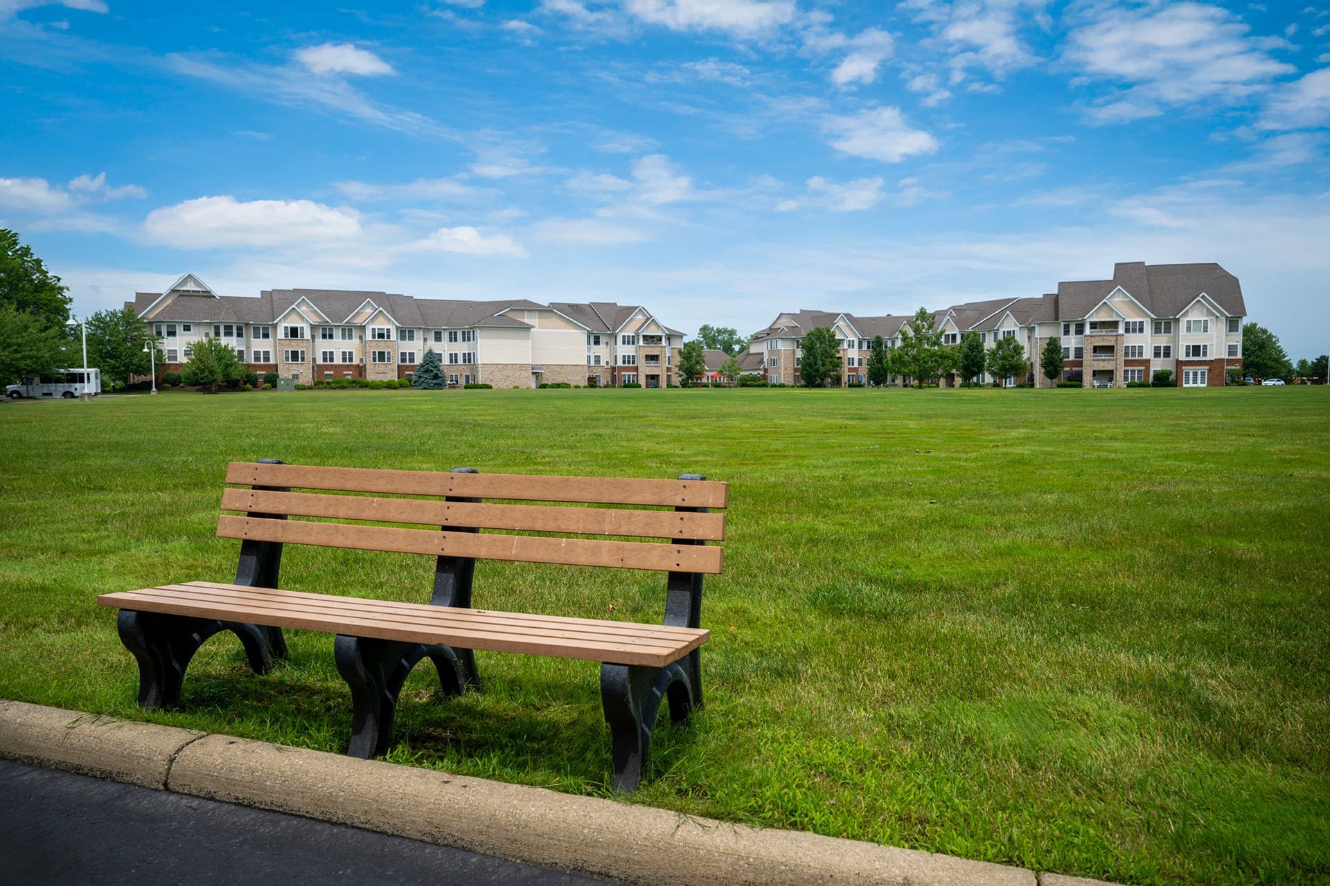 A brown park bench sits on a grassy field in front of a multi-story apartment complex under a blue, cloudy sky.