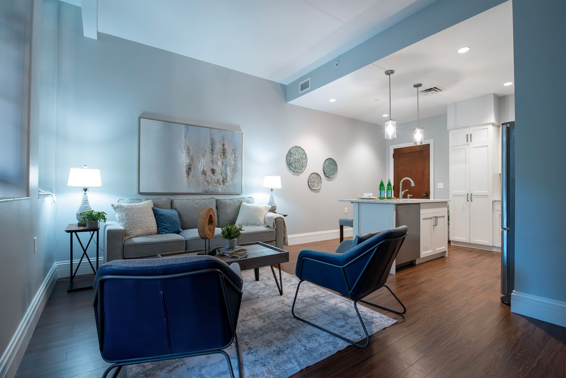 Bright open living room with blue chairs, sofa, wall art, and a kitchen island in the background