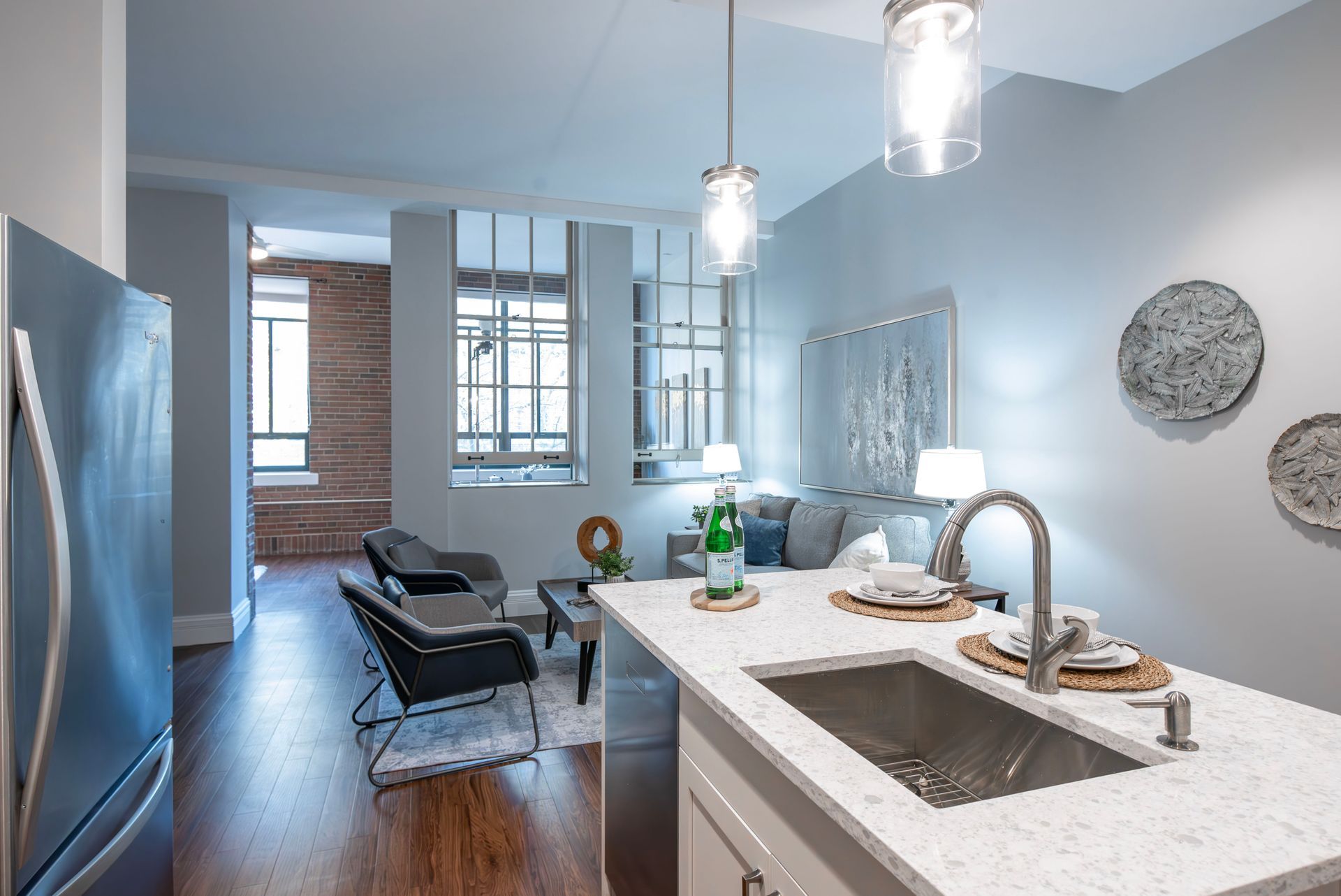 Modern kitchen with white island, pendant lights, and a view into a living area with brick wall and large windows
