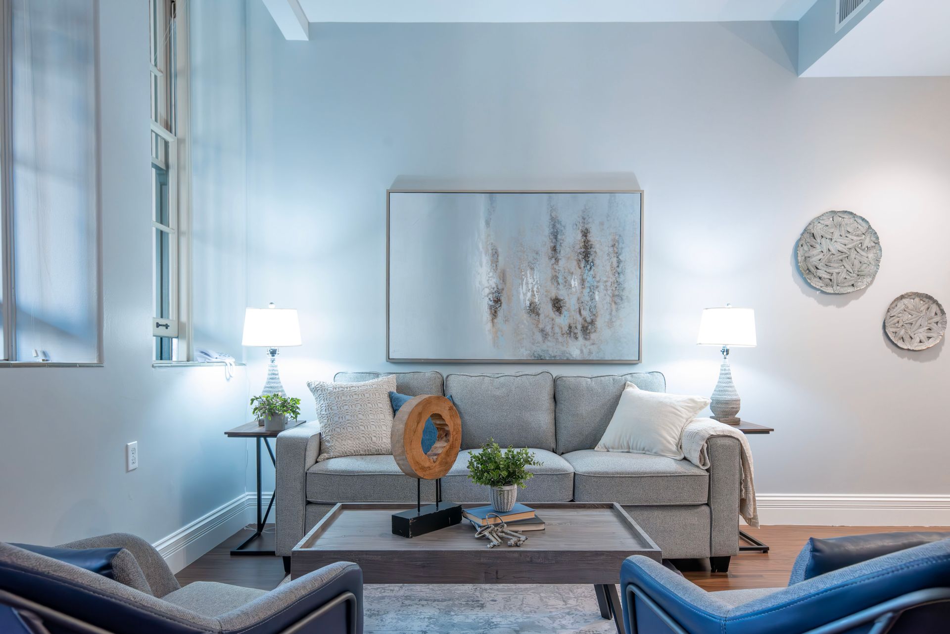Bright living room with a gray sofa, blue chairs, lamp, and abstract wall art