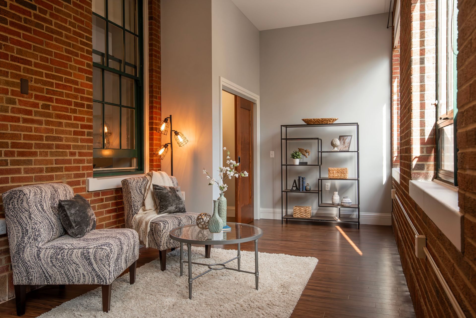 A cozy seating area with two patterned armchairs, a glass coffee table, a shag rug, and a metal shelf against brick walls.