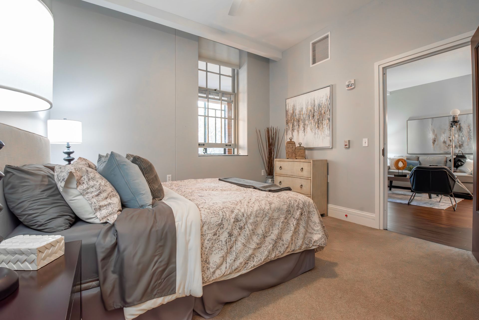 Cozy beige bedroom with a bed, window, lamp, and full-length mirror reflecting an adjacent room.