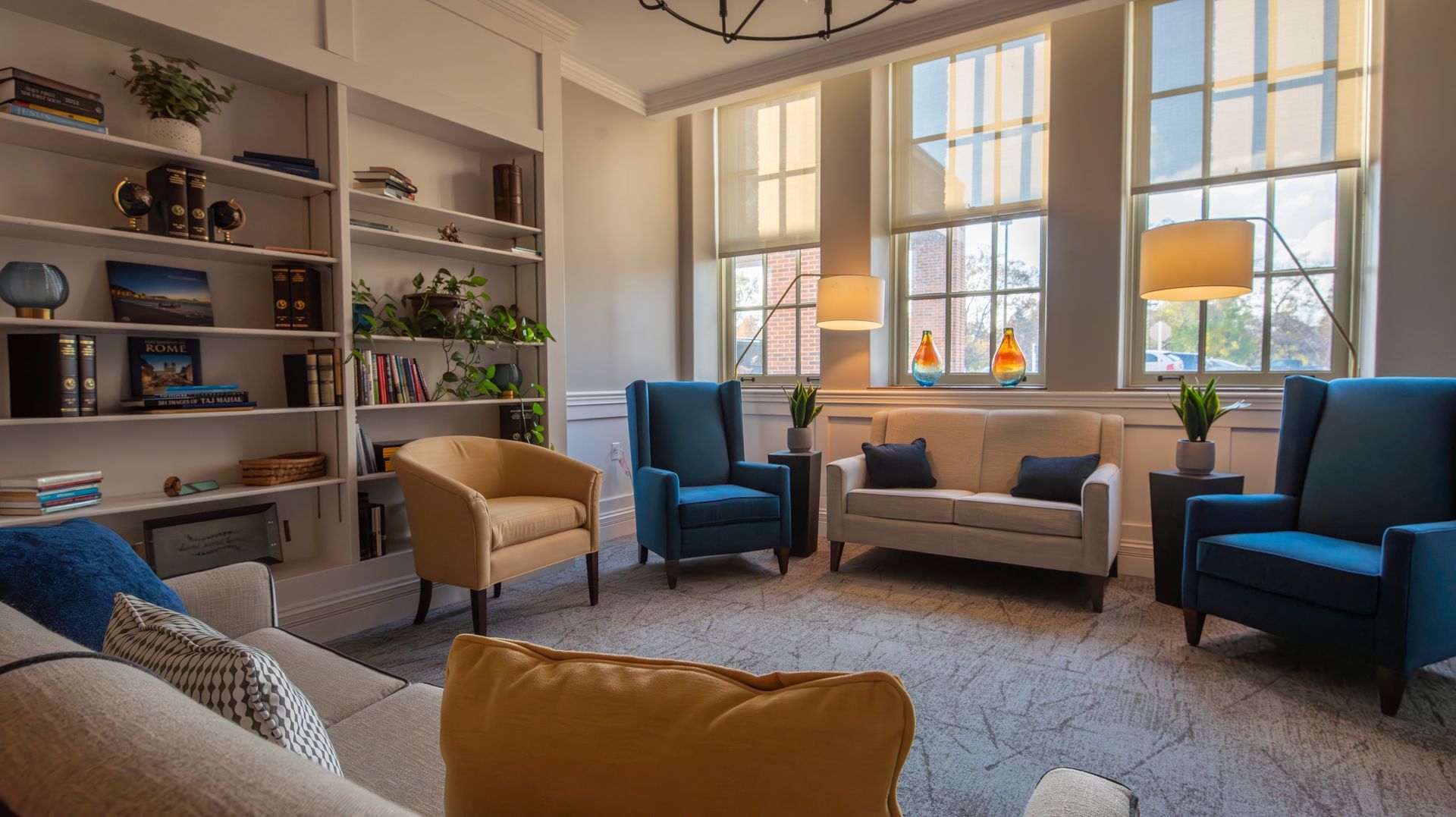 A cozy library seating area with white built-in shelves, a beige sofa, blue armchairs, and yellow chairs on a rug.