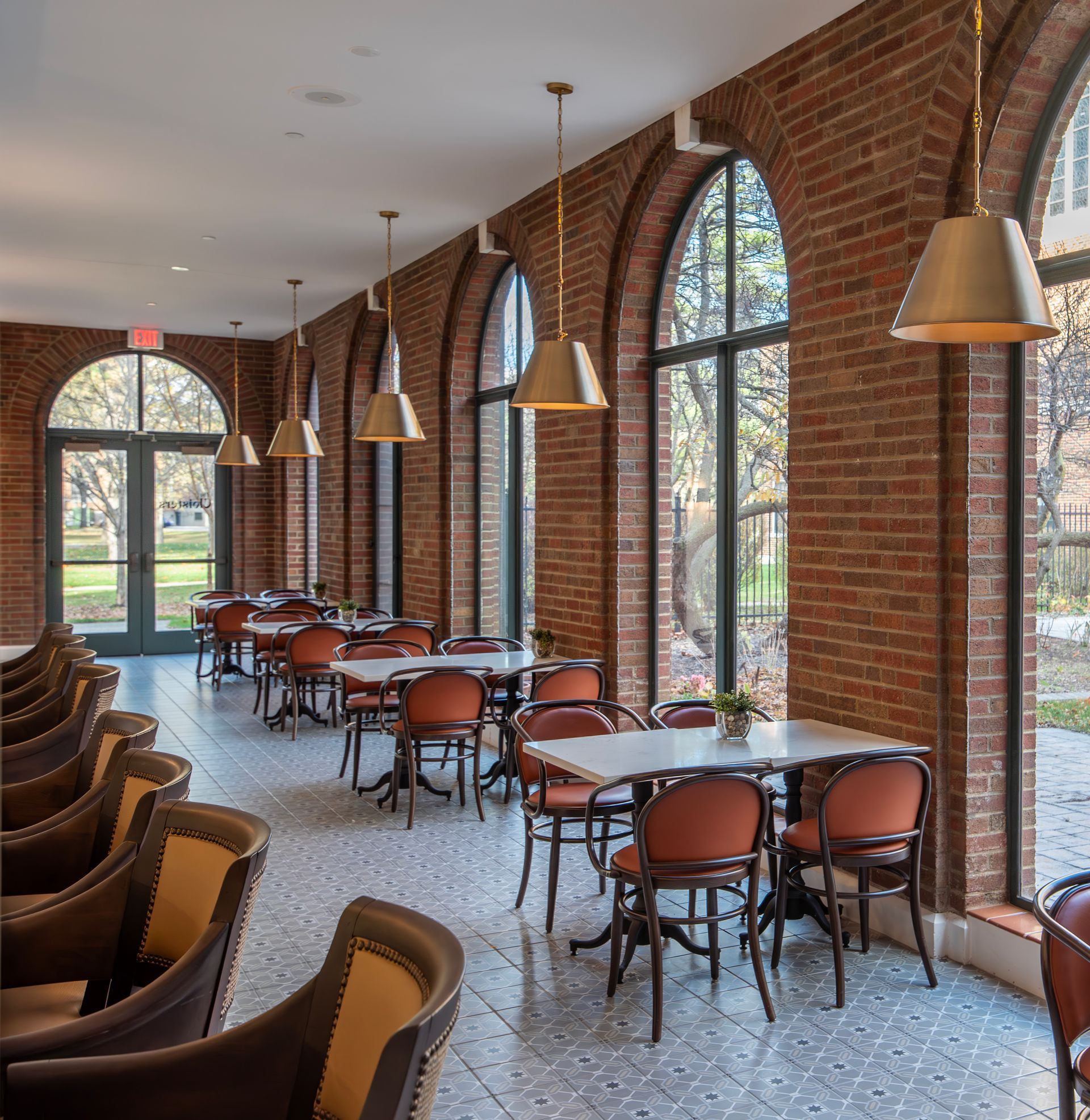 Empty dining area with brick walls, arched windows, modern pendant lights, and tables with chairs set on patterned floors.