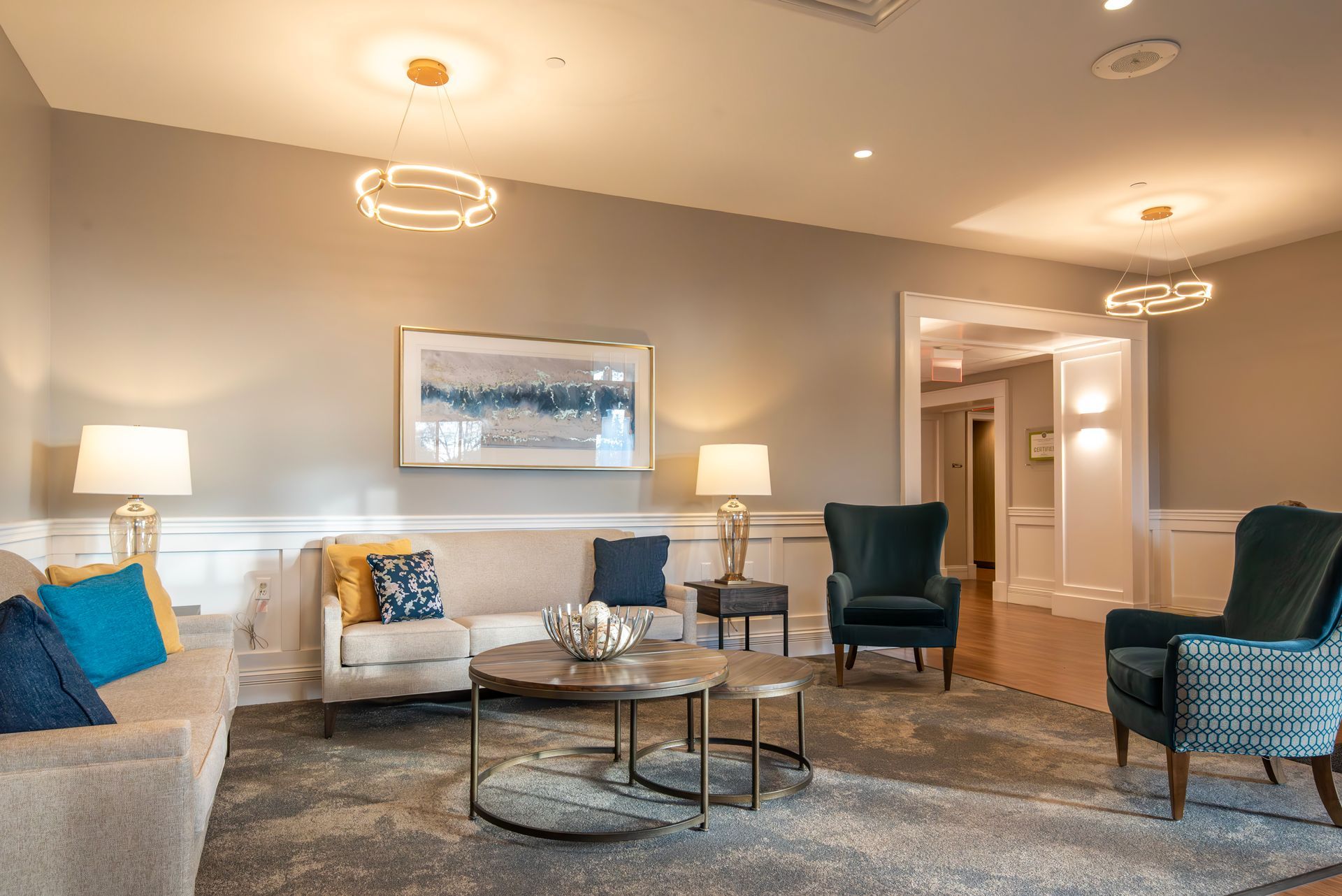 A well-lit sitting area with a light-colored sofa, patterned blue armchairs, round coffee tables, and wall-mounted art.