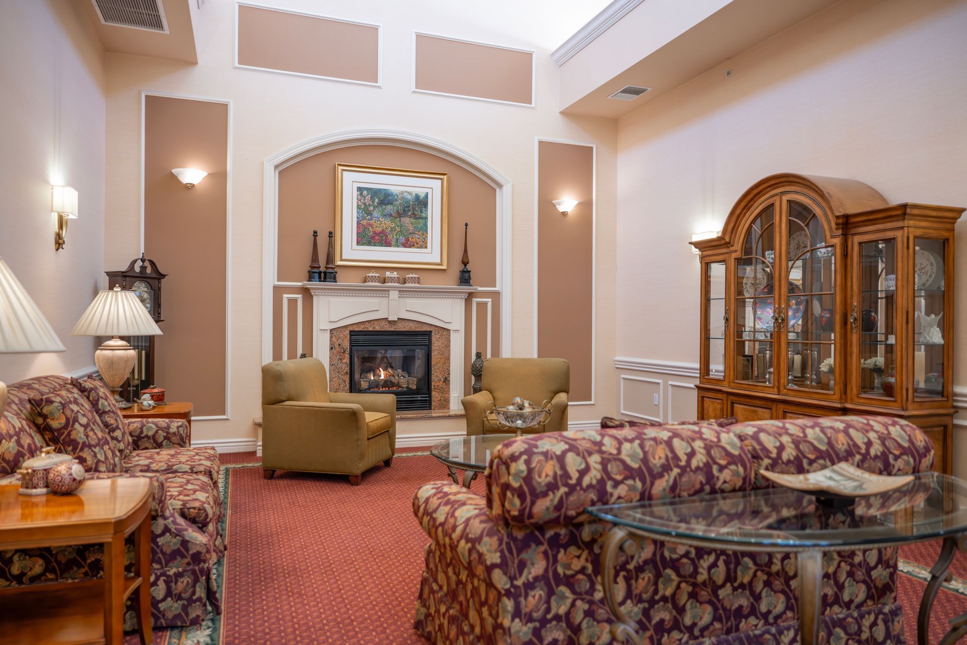A formal living room with two patterned sofas, two tan armchairs, a fireplace, and a large wooden display cabinet.