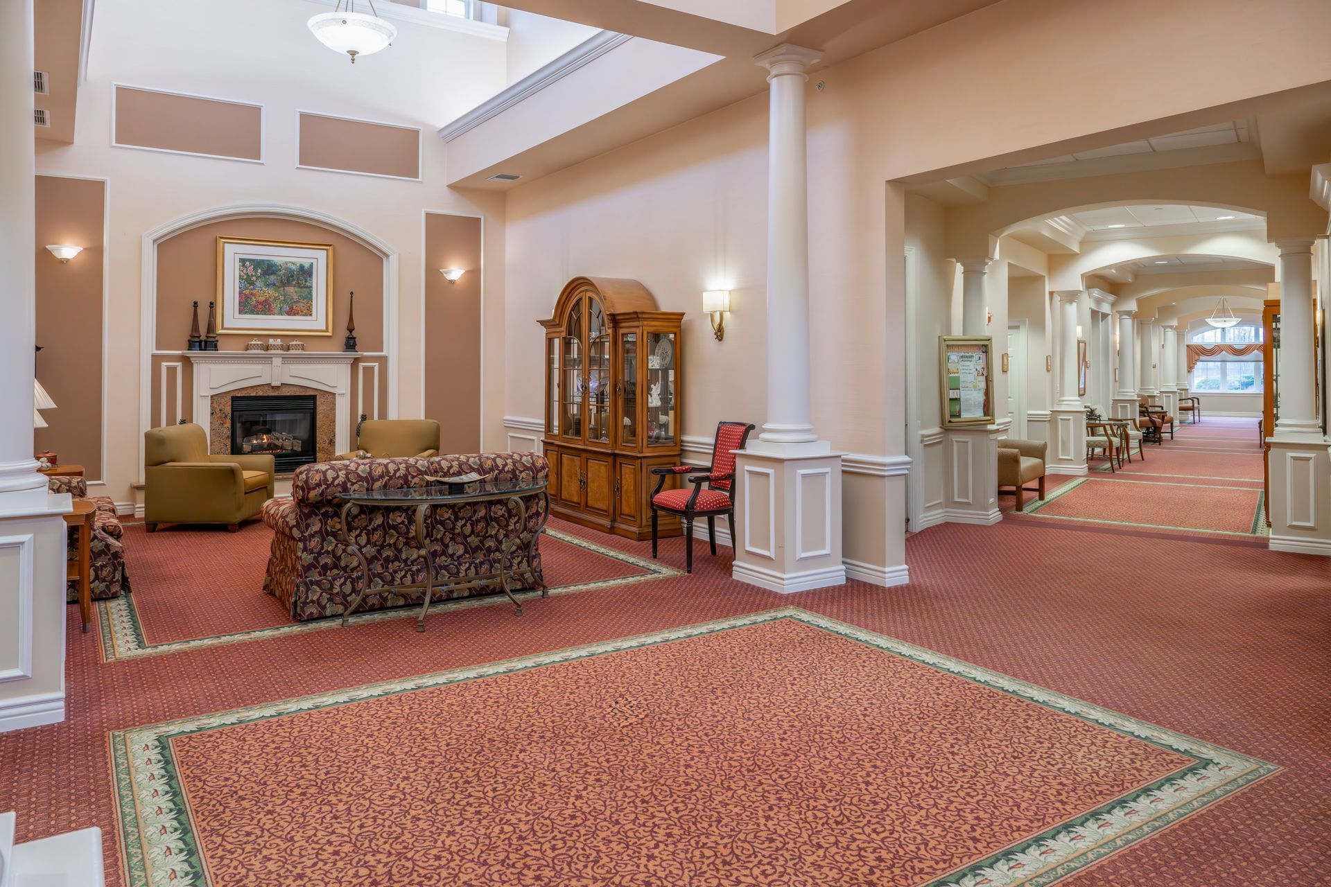 A spacious, well-lit lounge area with patterned carpeting, a fireplace, comfortable seating, and an arched hallway view.