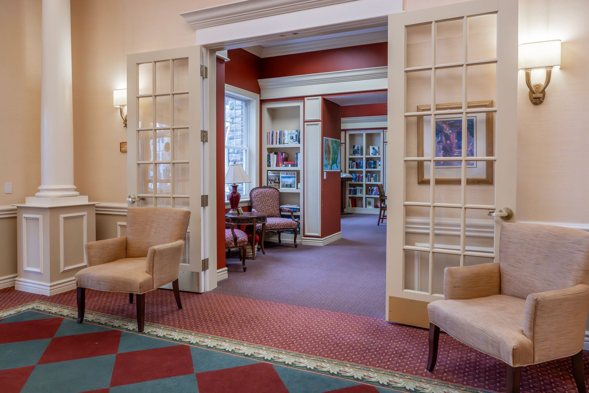 Two upholstered armchairs face a doorway leading to a library room with bookshelves, red walls, and patterned carpet.