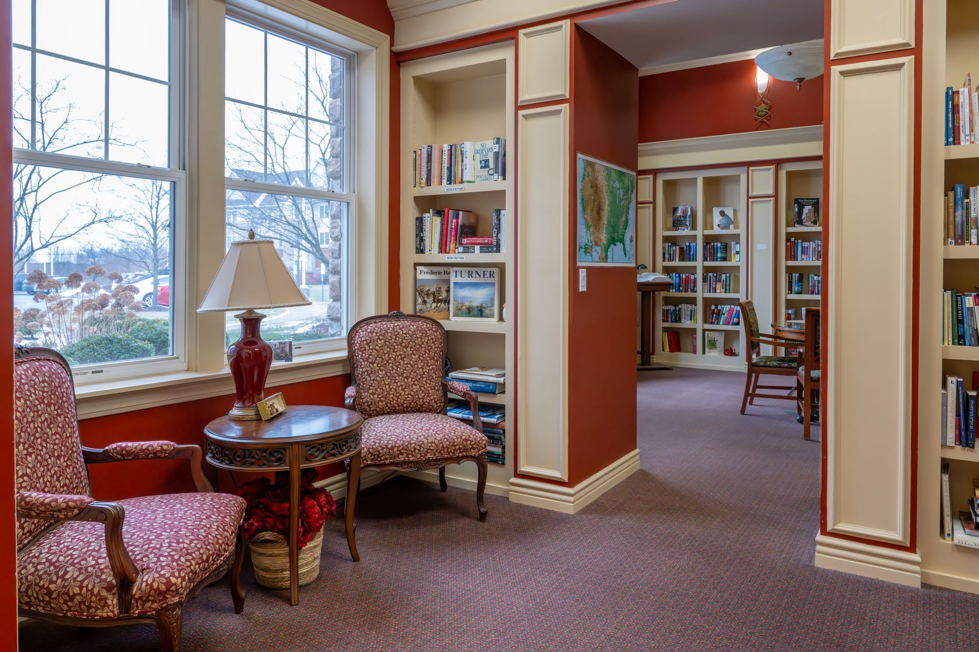 Two patterned armchairs sit by a window next to a lamp-topped side table in a library with red walls and built-in shelves.