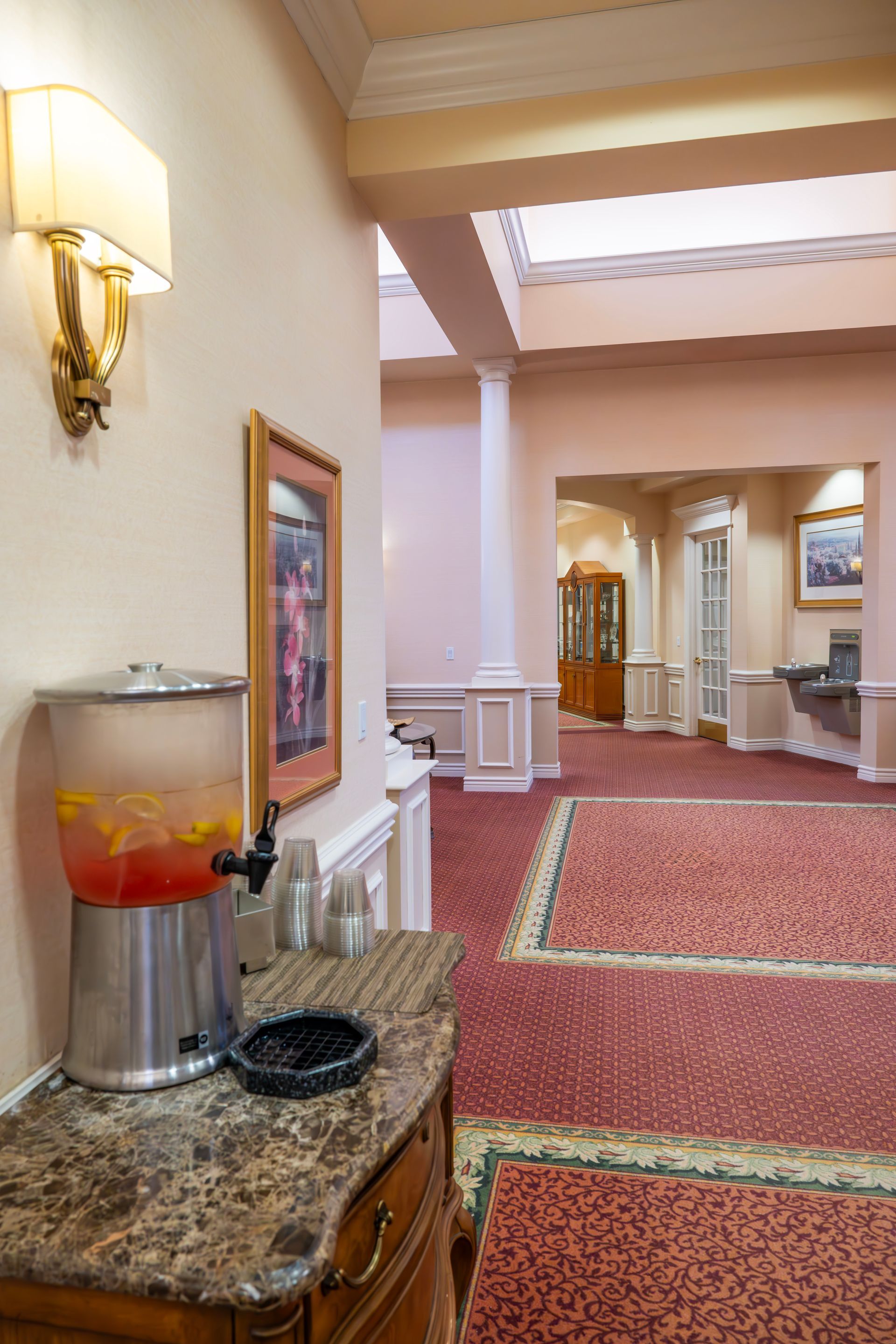 A beverage dispenser with fruit-infused water sits on a marble-topped counter in a hallway with red patterned carpeting.