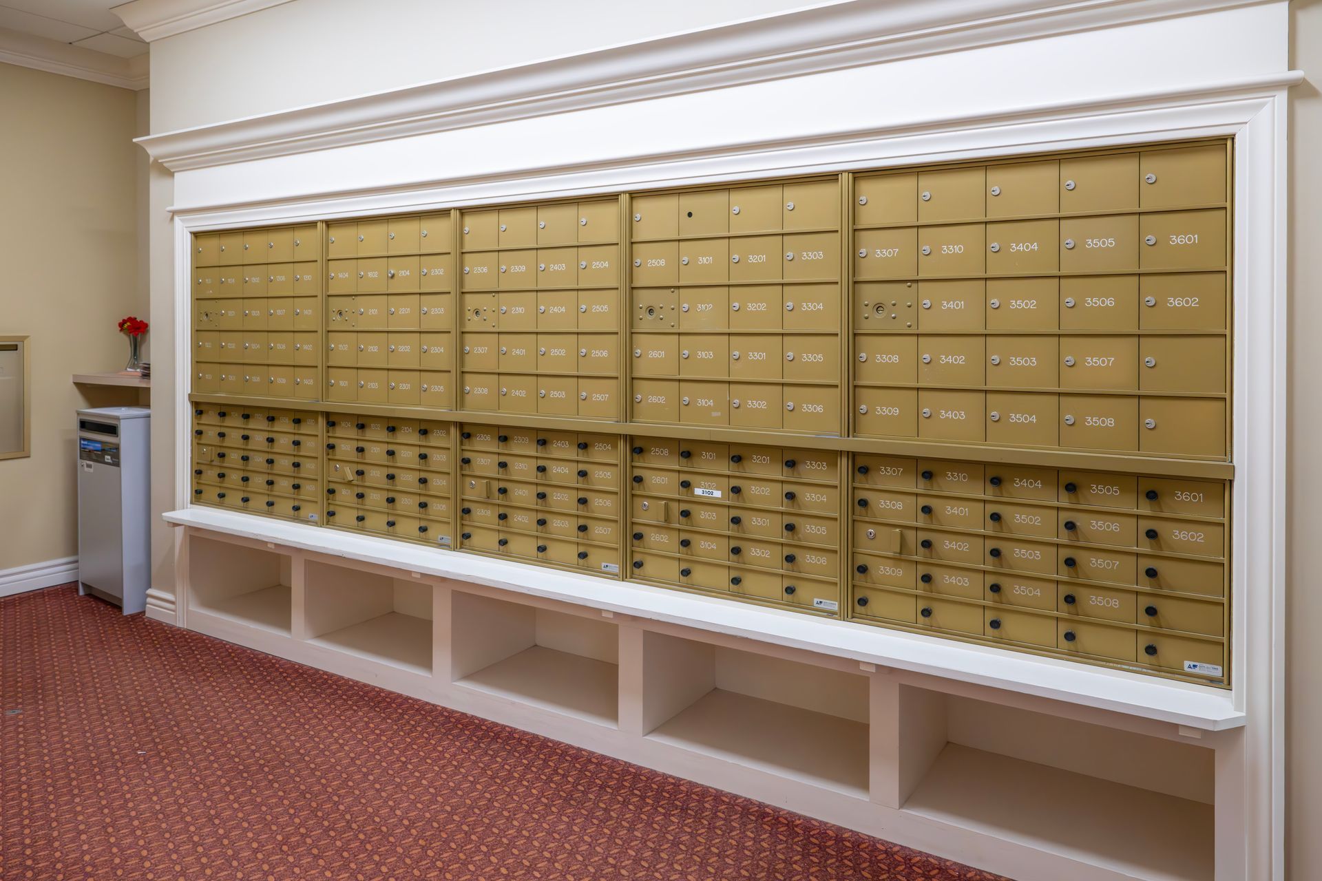 A wall of metal mailboxes with numbered slots, set in a room with patterned carpet and built-in white shelving below.