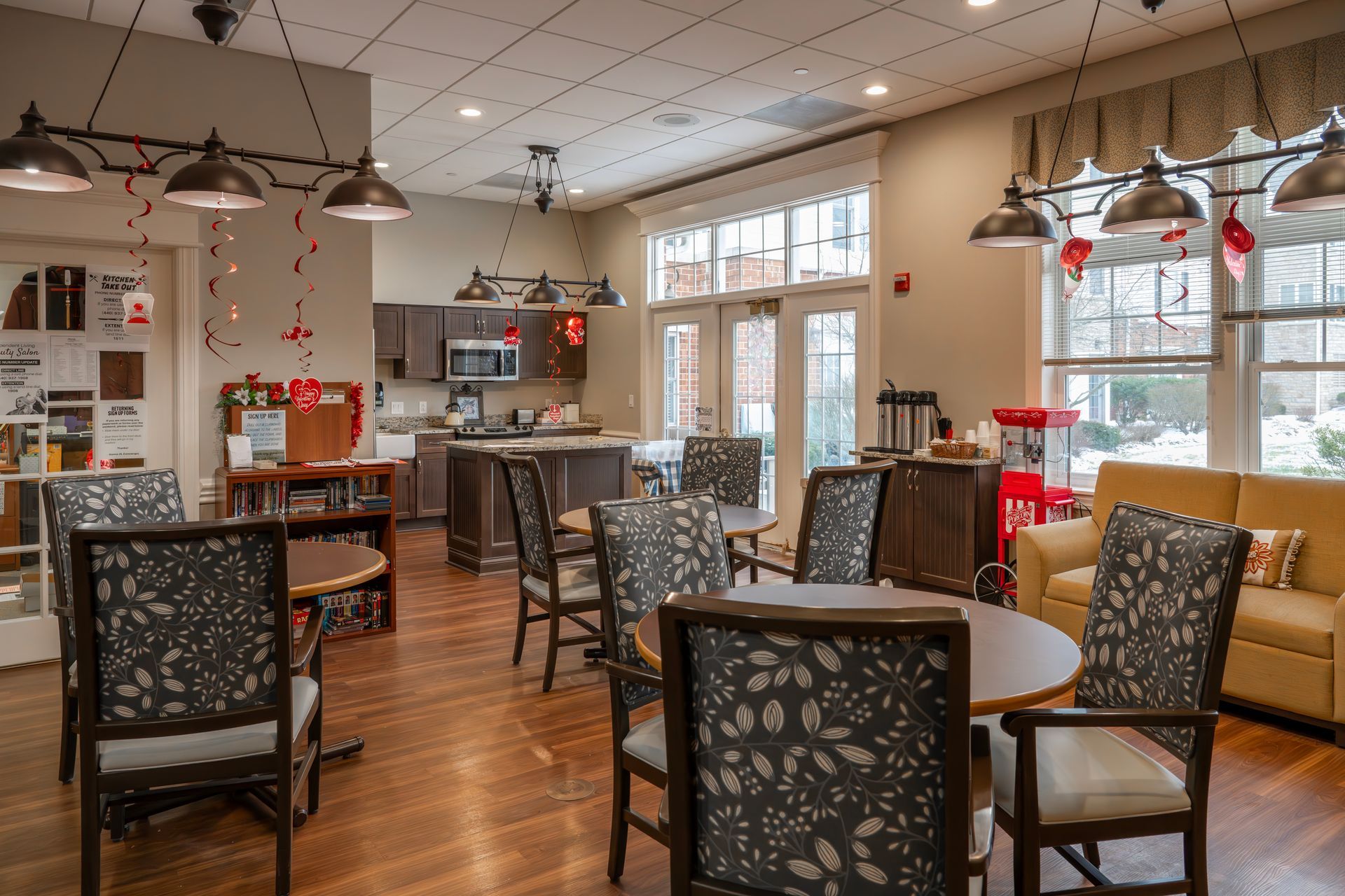 A community lounge with round tables, patterned chairs, a kitchen area, and a yellow sofa.