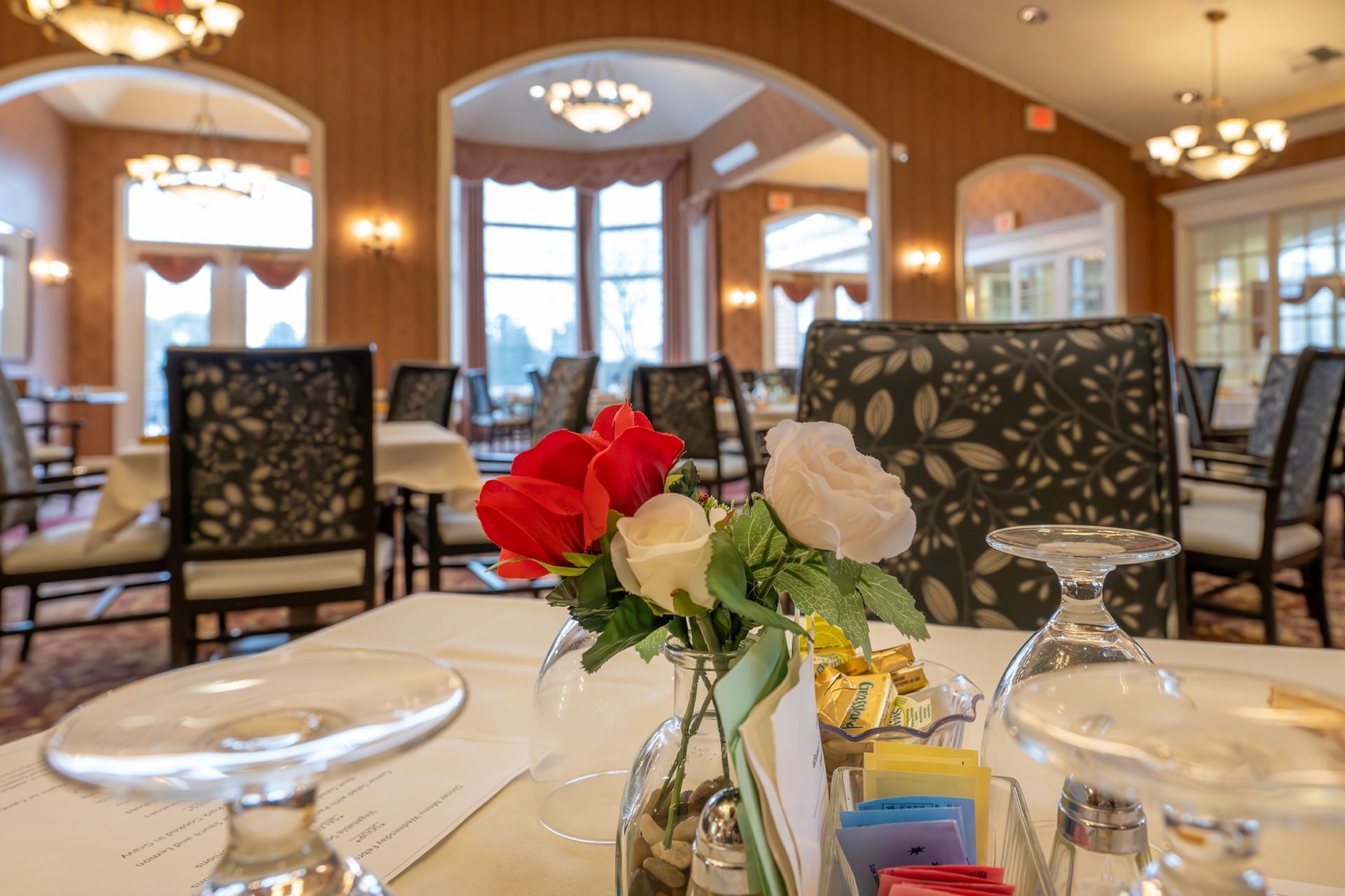 A close-up of a small flower vase with red and white roses on a dining table inside an elegant, sunlit restaurant.