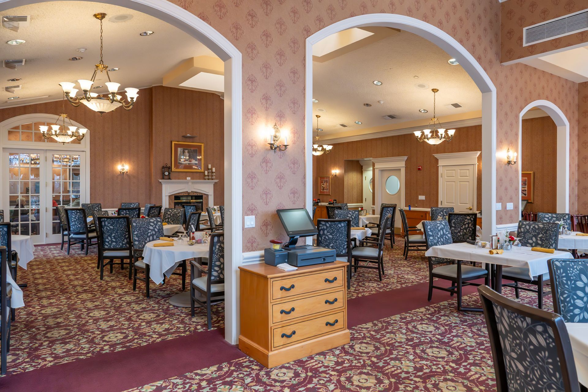 A dining room with arched walls, patterned carpeting, chandeliers, and tables set for service in a warm, formal space.