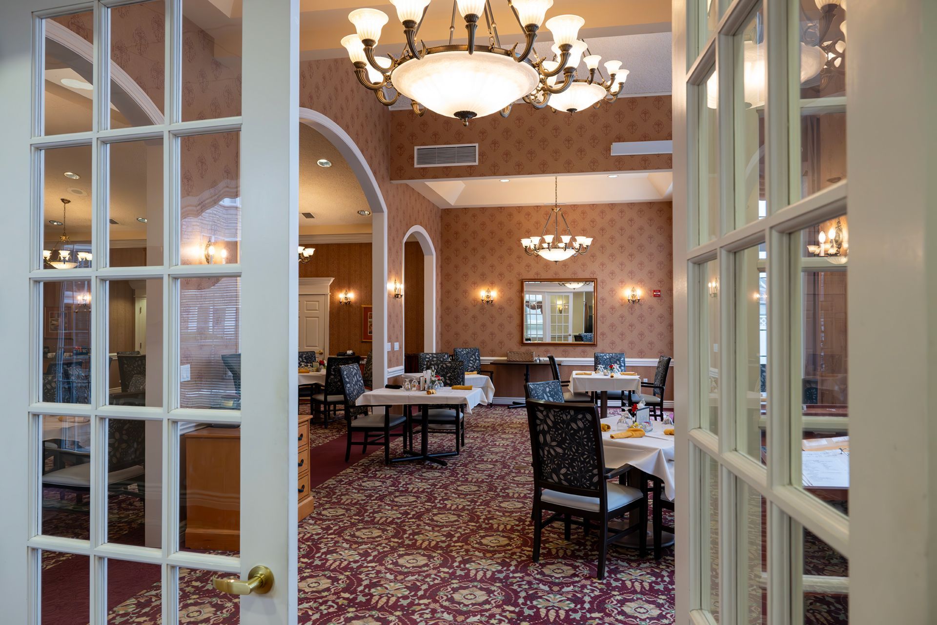 Dining room with tables, patterned carpet, and chandeliers, viewed through open French doors.