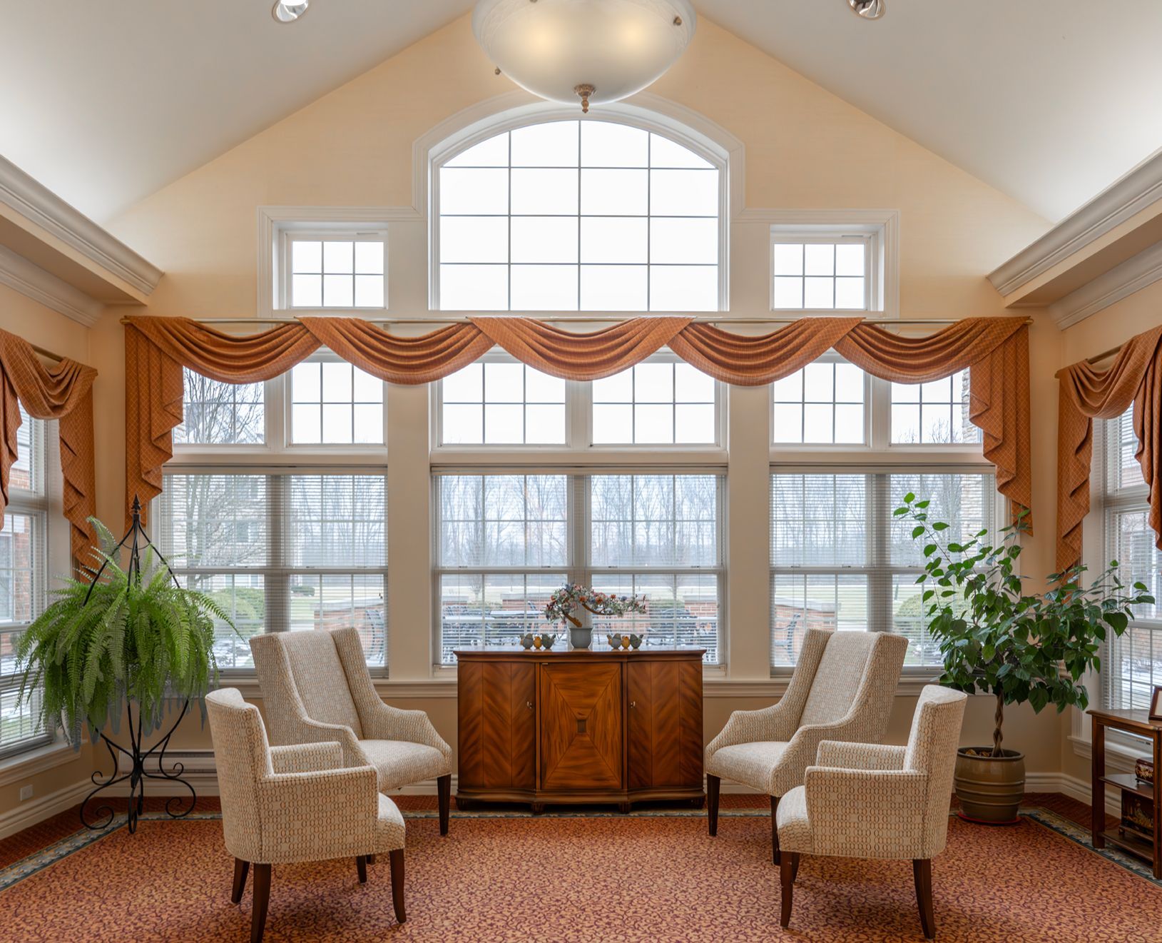 A sunlit seating area with beige armchairs, a wooden cabinet, and large windows with orange valances over a patterned rug.