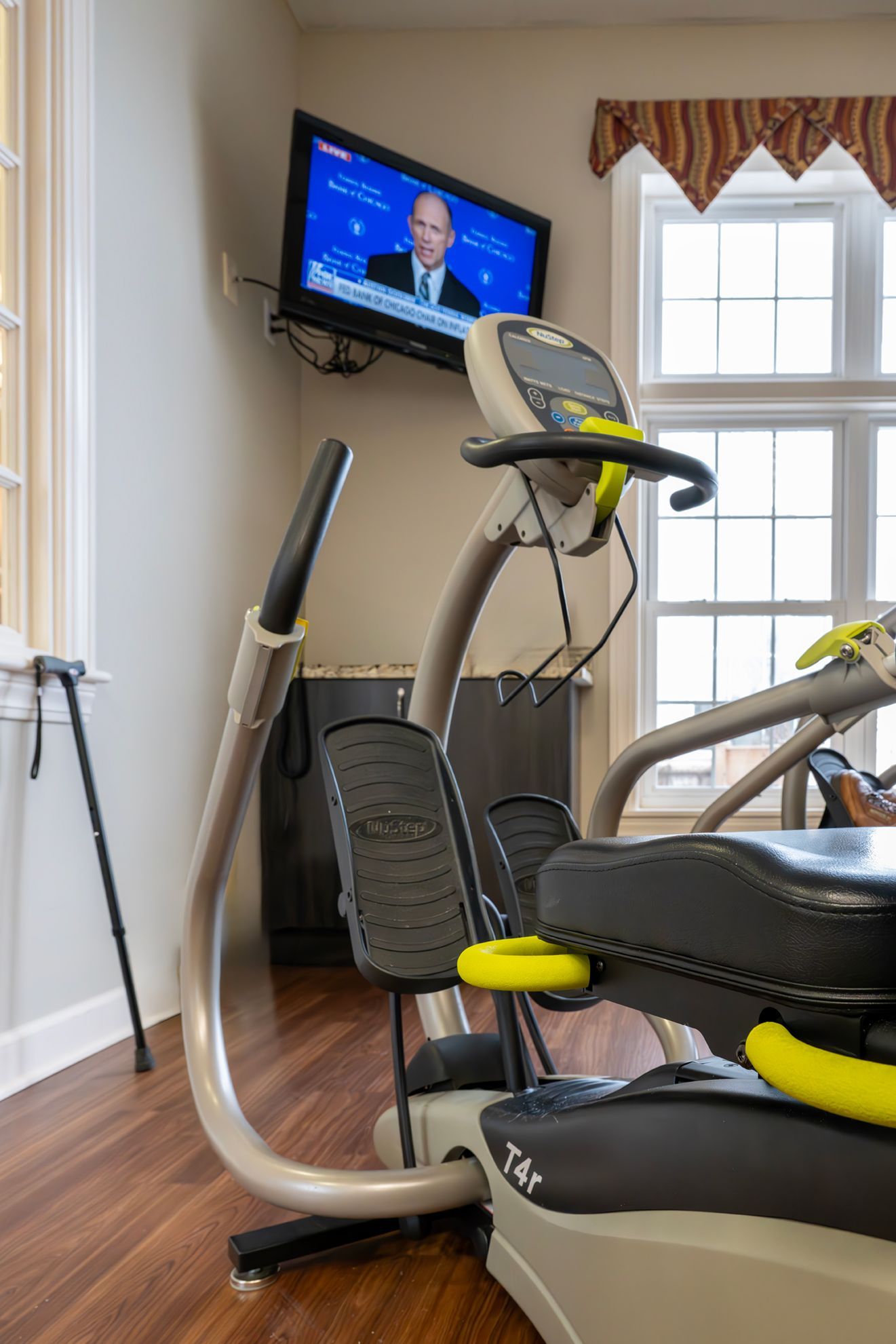 A gray and black elliptical machine in a room with hardwood floors, a wall-mounted TV, and a walking cane in the corner.