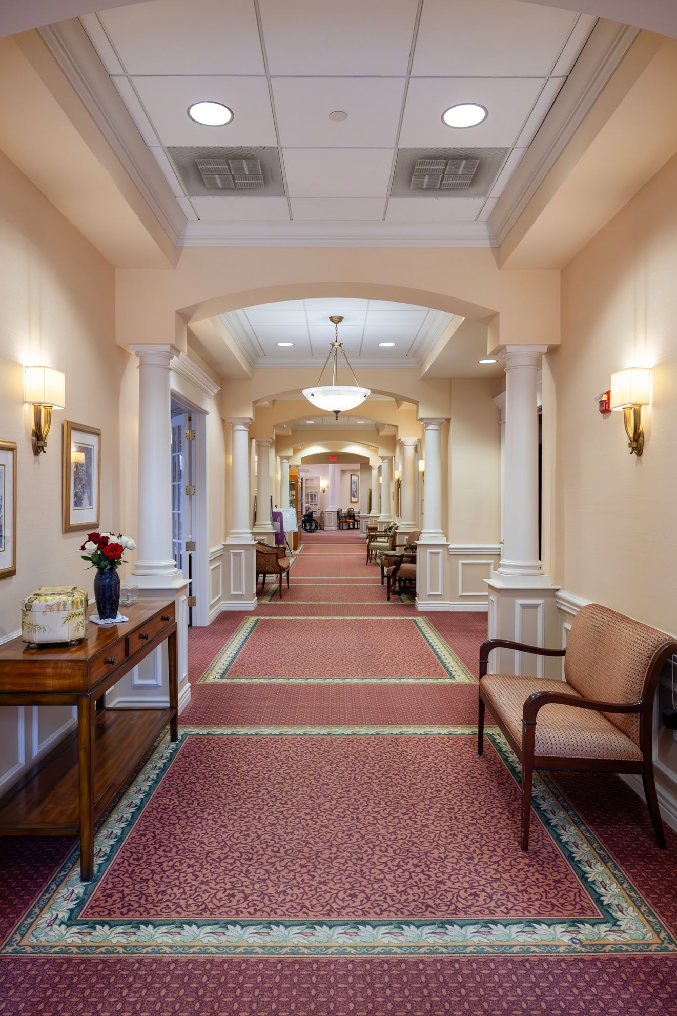 A long, carpeted hallway with columns, decorative wall sconces, a wooden console table, and a settee.