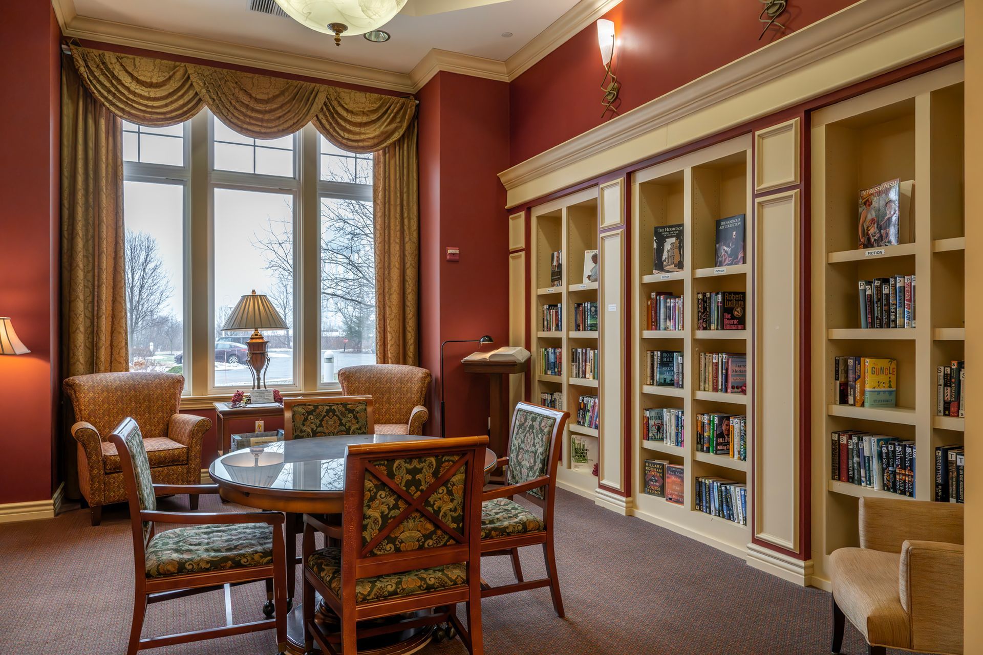 A quiet library room with red walls, bookshelves, a round table with chairs, and armchairs near a large window.