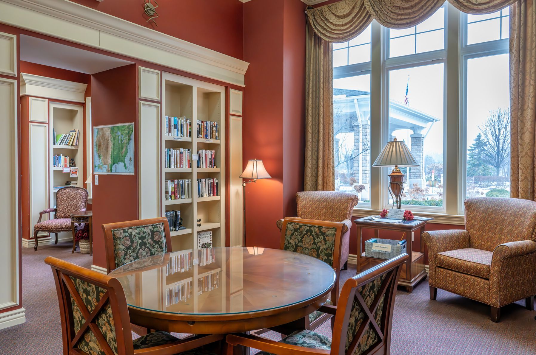 A library seating area with a glass-topped table, patterned armchairs, bookshelves, and a large window with draped curtains.