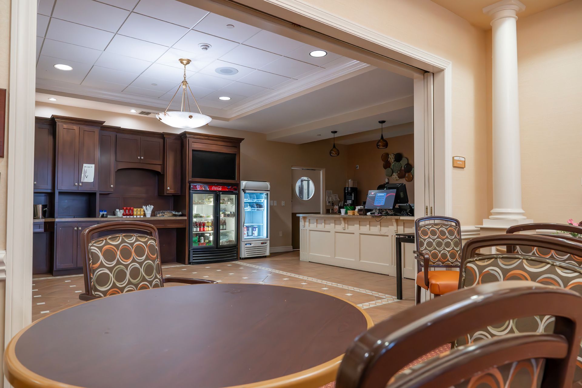 A break area with round tables and chairs, featuring a kitchen corner with dark cabinets and a glass-front refrigerator.