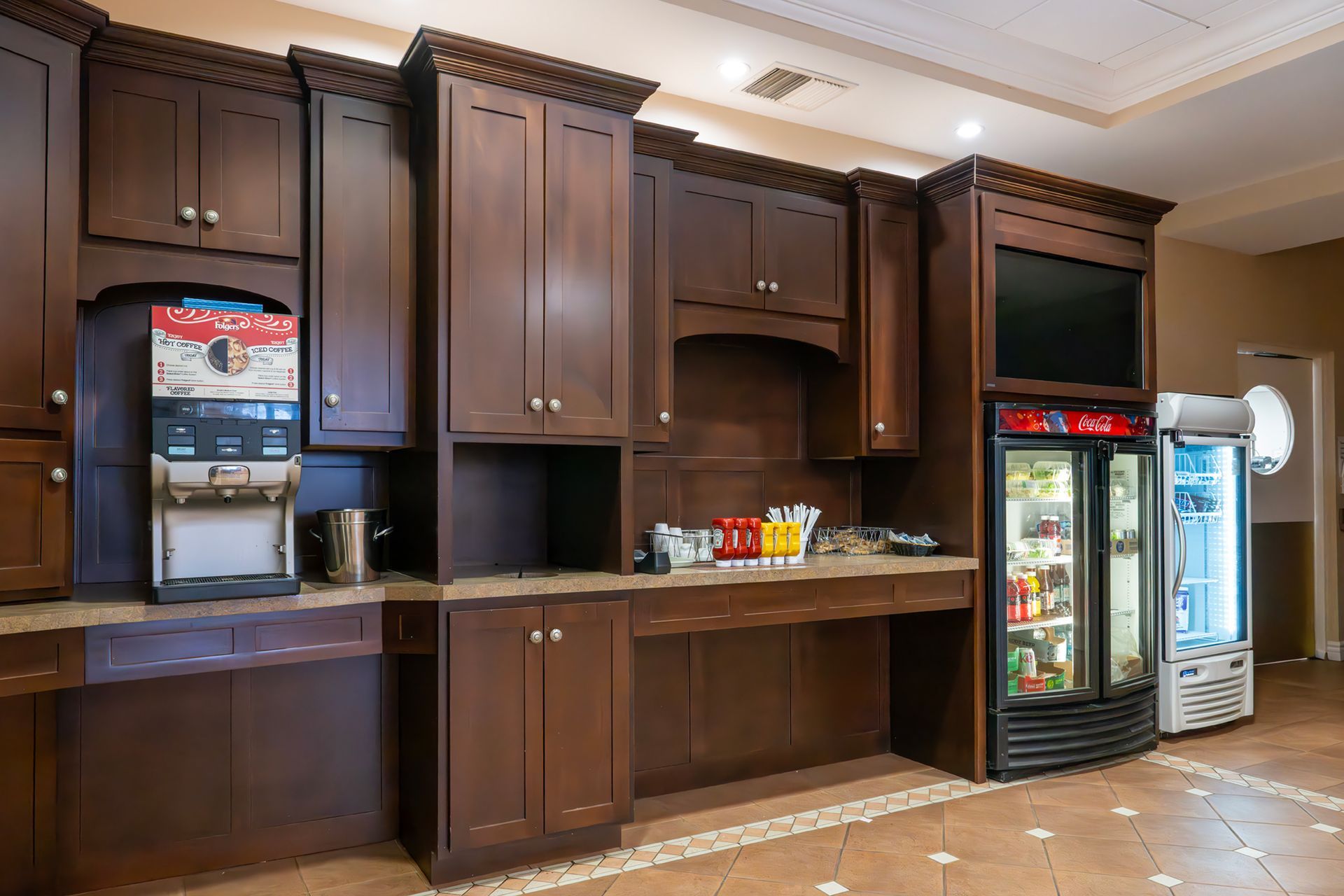 A hotel beverage station with a coffee machine, condiments, and two glass-fronted refrigerators in dark wood cabinetry.